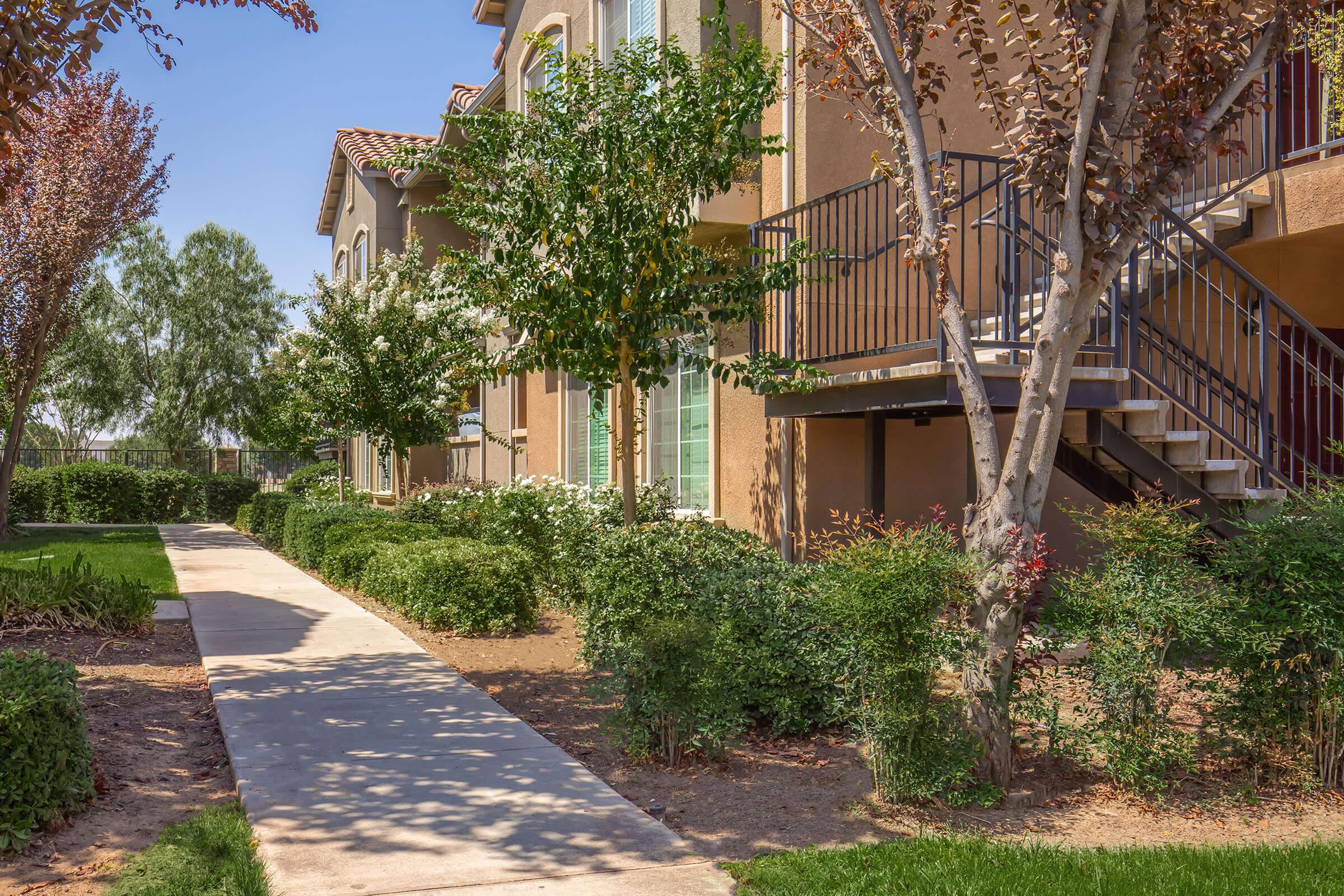 A view of a landscaped pathway leading to a multilevel residential building. The area features green shrubs and trees with some colorful foliage, providing a pleasant outdoor environment. The building has balconies and a staircase, set against a clear blue sky.
