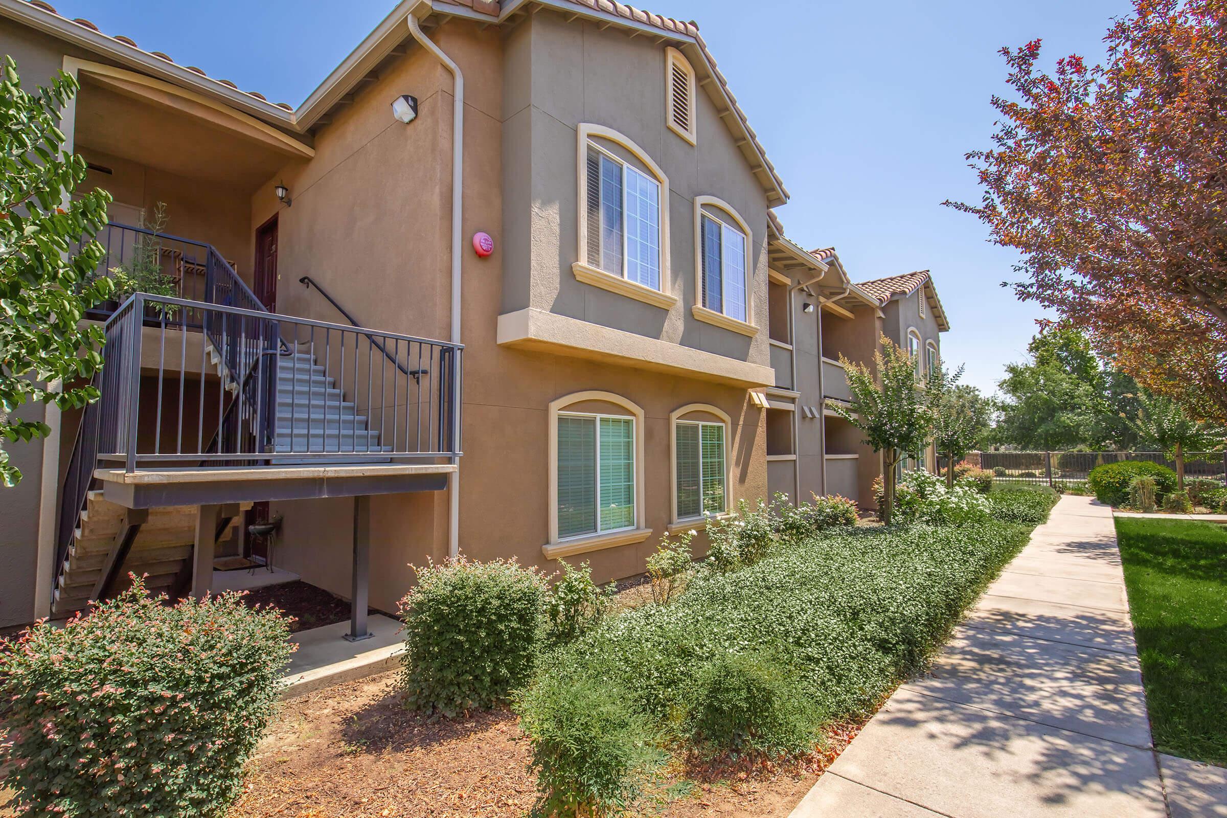 A well-maintained multi-unit residential building with a staircase leading to upper levels. The exterior features warm beige stucco and large windows. Lush green shrubs line the walkway, which leads to grassy areas in the background. Trees provide shade, and the sky is clear and blue, indicating a sunny day.