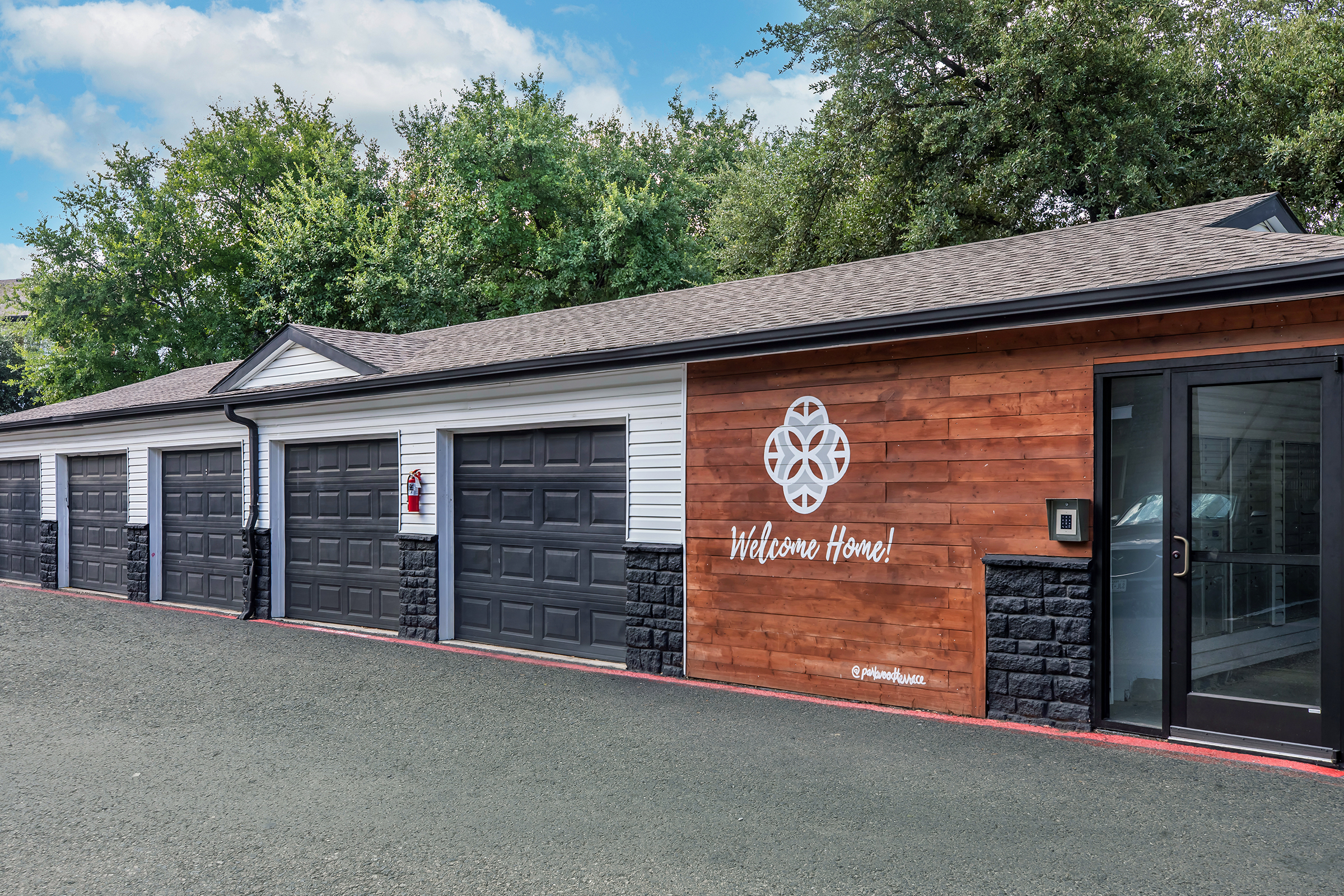 A storage facility with a row of garage doors, featuring a welcoming sign on one side that reads "Welcome Home!" The building has a mix of white and wooden siding, and there are trees in the background. A security keypad is visible near the entrance. The ground is paved with dark materials.