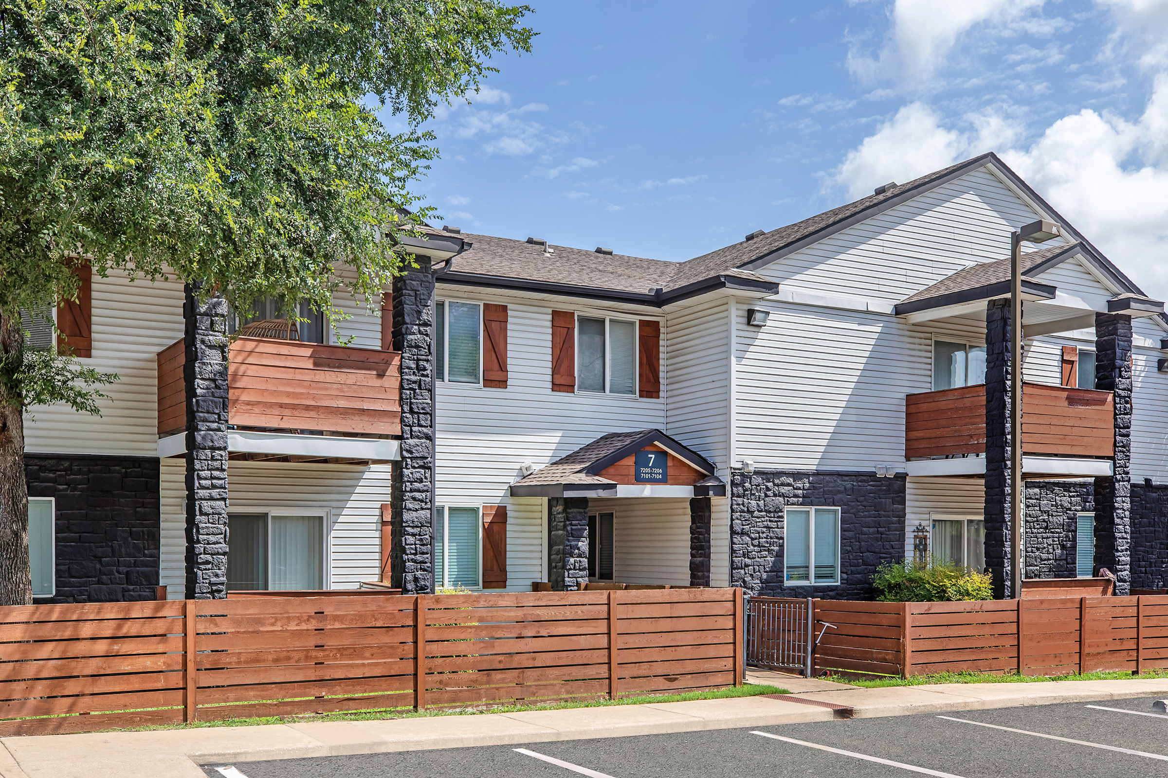 A two-story residential building with white siding and dark stone accents. It features wooden balconies on the upper floor and a fenced area in front. The area is landscaped with trees and shrubs, under a partly cloudy sky. Parking spaces are visible in the foreground.