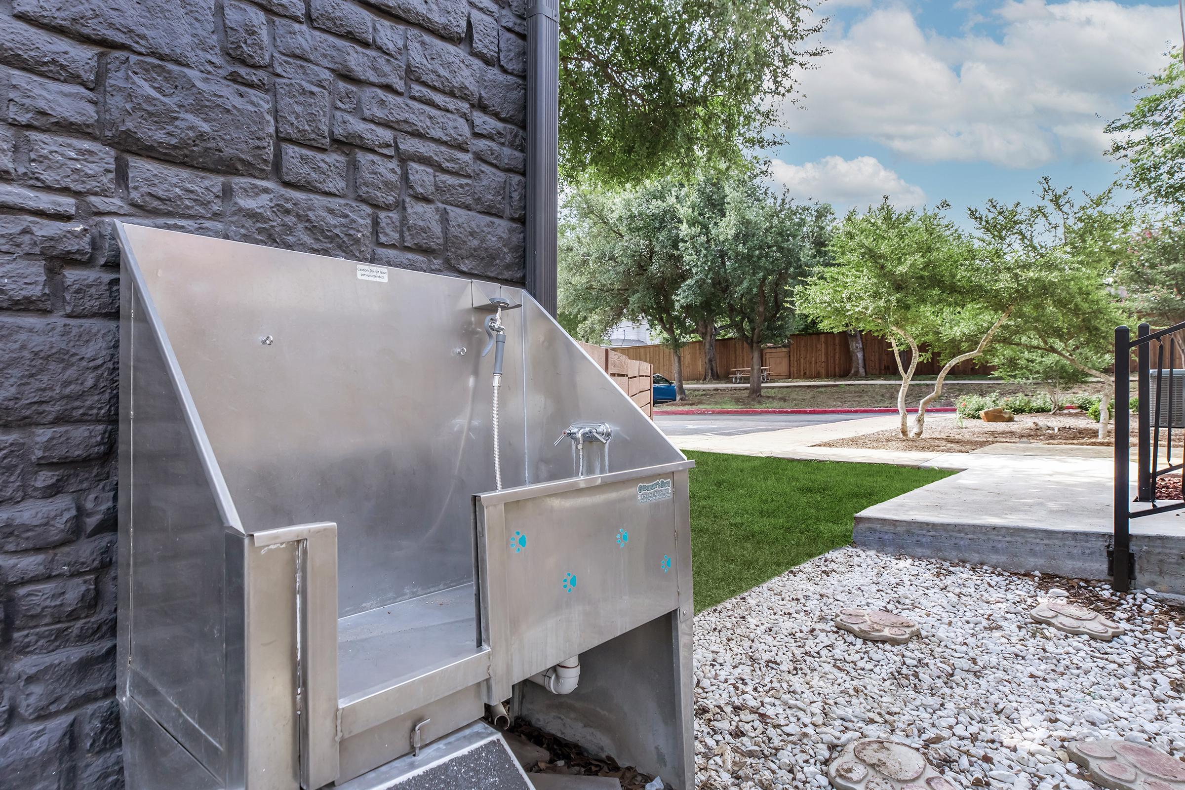 A stainless steel outdoor utility sink positioned against a textured gray stone wall. The area features a landscaped yard with green grass, trees, and a pathway. The sky is partly cloudy, adding a serene atmosphere to the outdoor space.