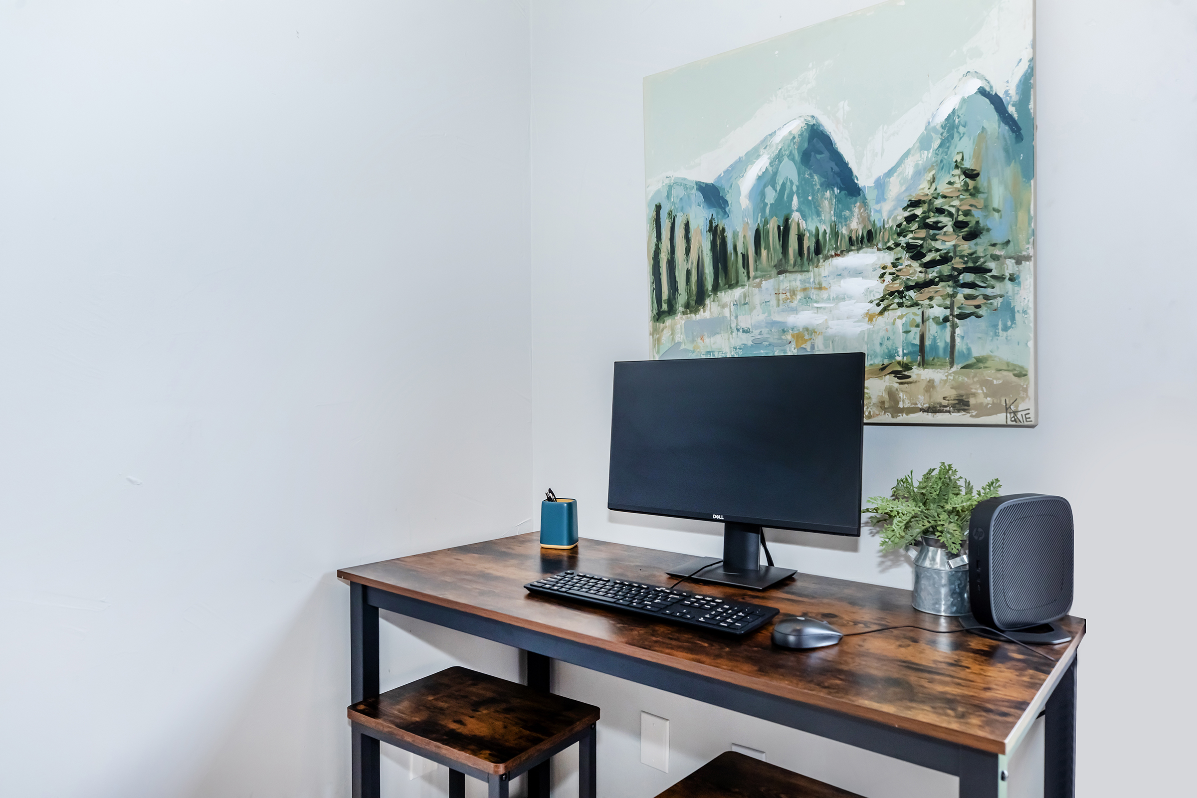 A modern workspace featuring a wooden desk with a computer monitor, keyboard, and mouse. A small blue container and a potted plant are on the desk. The wall has a landscape painting depicting mountains and trees, adding a serene touch to the minimalist setup.