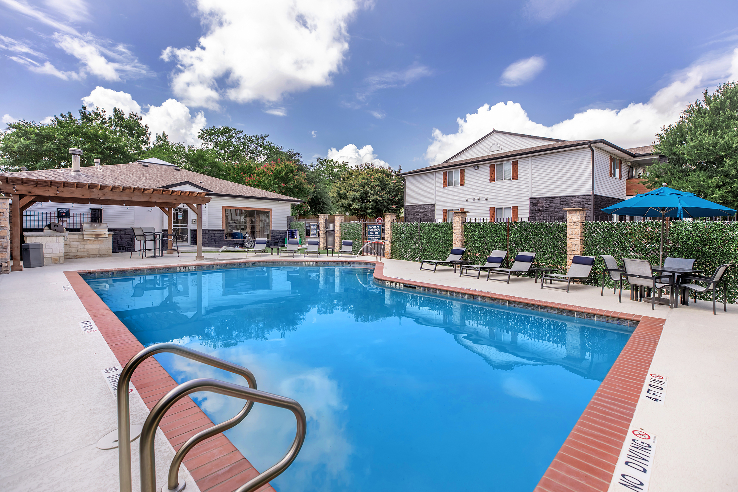A clear blue swimming pool surrounded by lounge chairs and umbrellas, with a shaded pergola on one side. Lush greenery and two buildings are visible in the background under a partly cloudy sky. The scene conveys a relaxing outdoor space perfect for leisure activities.