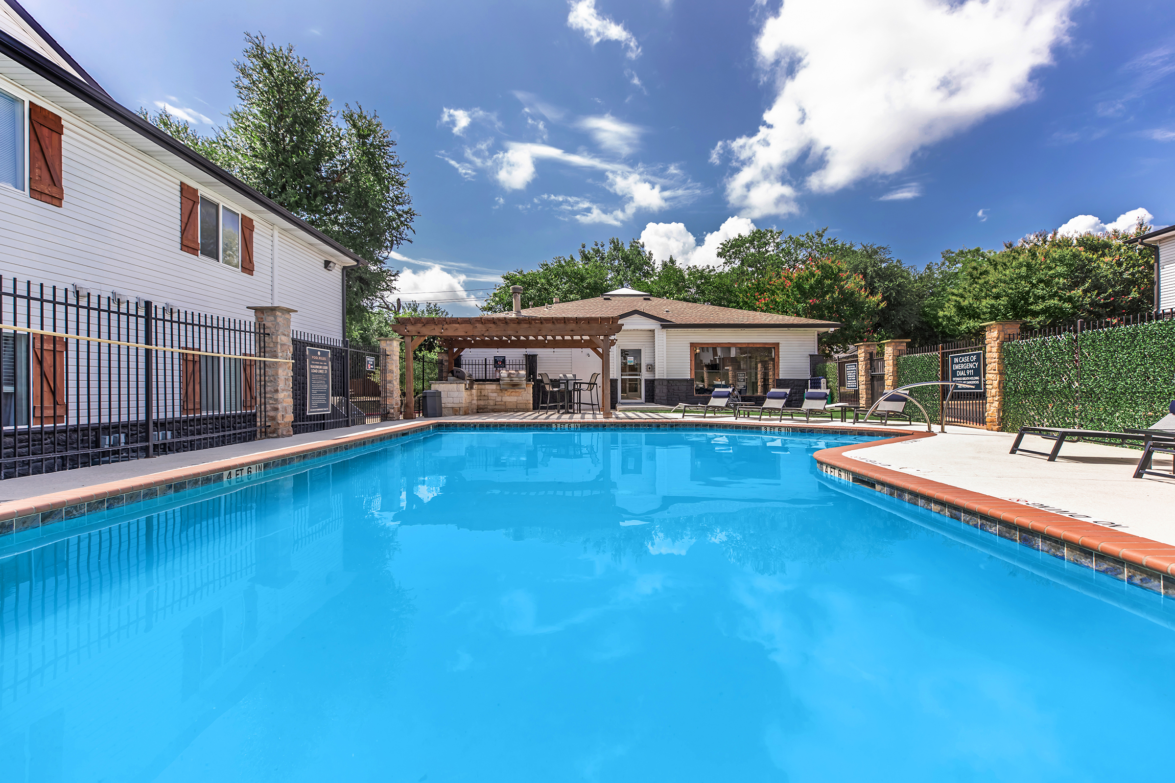 A clear blue swimming pool surrounded by lounge chairs, with a white house and outdoor pergola in the background. The sky is bright with fluffy clouds, and lush greenery is visible around the property, creating a relaxing outdoor space for leisure and recreation.