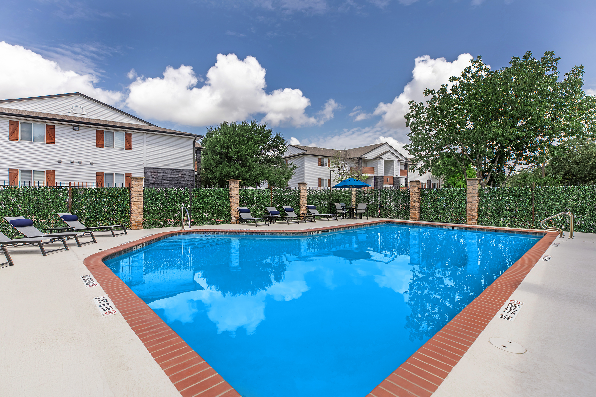 A vibrant blue swimming pool surrounded by lounge chairs, with green hedges and a few trees in the background. In the distance, there are two buildings with white exteriors. The sky is partly cloudy, creating a bright and inviting atmosphere.