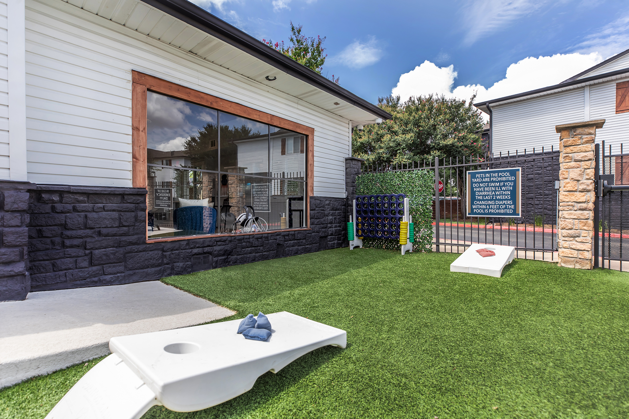 Exterior view of a recreational area featuring a well-maintained green lawn, a bean bag toss game, and a fenced patio. A large window showcases an indoor area, and there are informational signs nearby. The setting is bright and inviting, ideal for leisure activities.