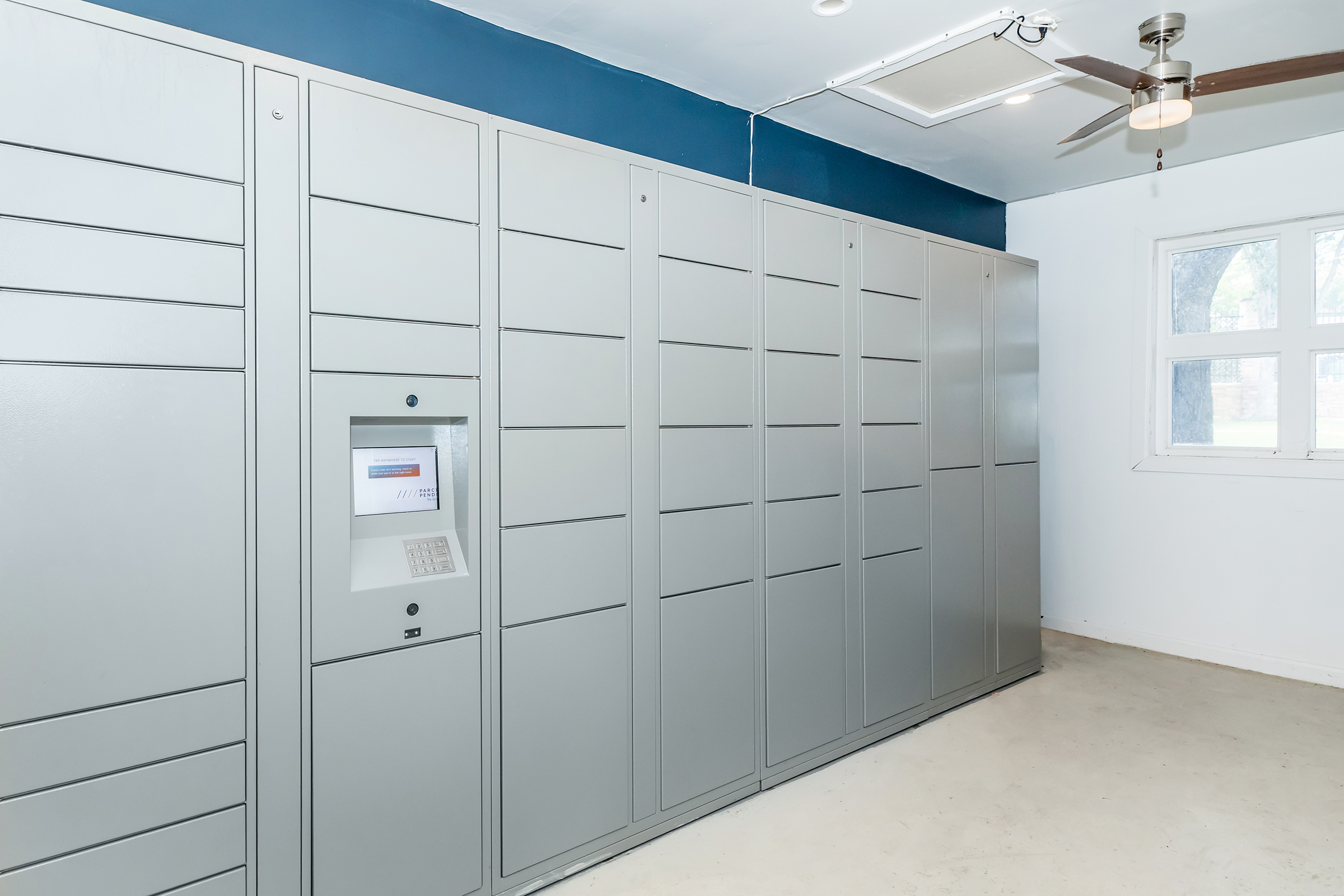 A modern interior space featuring a row of gray lockers against a blue wall. There is a digital package delivery kiosk at one end of the lockers. The floor is light-colored, and a ceiling fan is visible. A window allows natural light into the room.