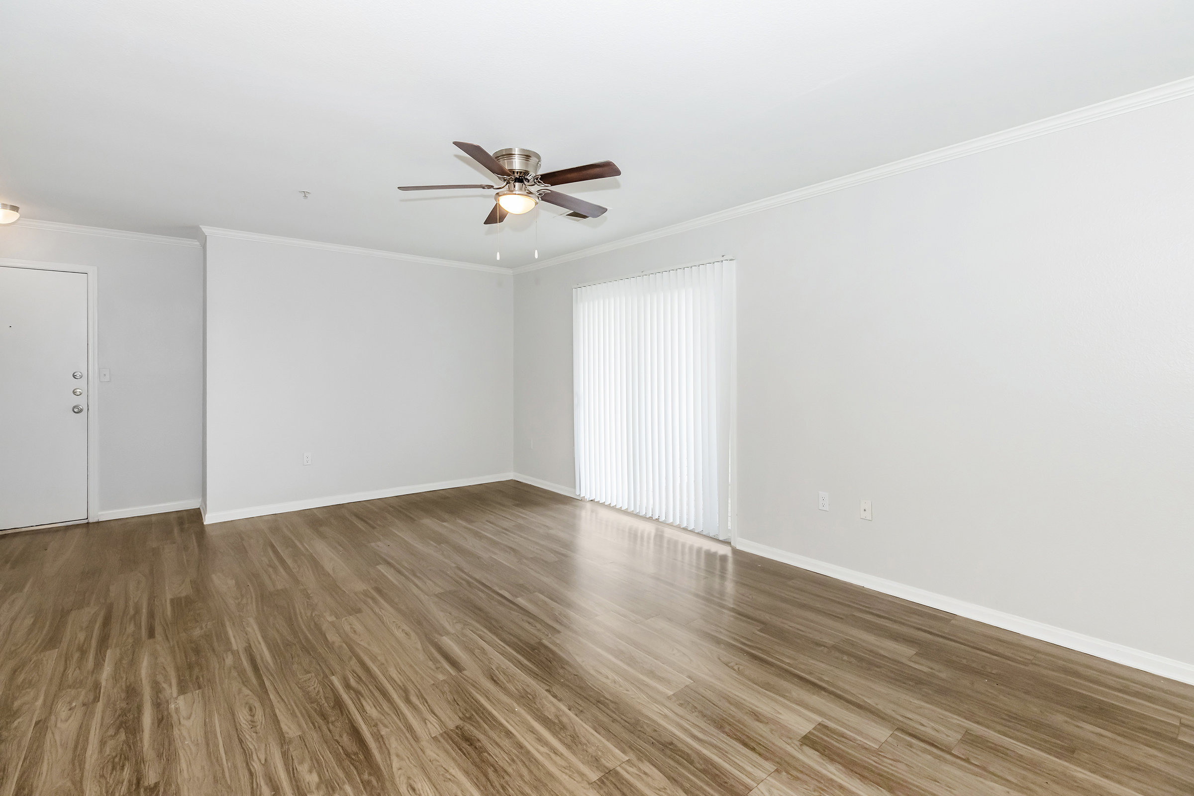 A spacious, empty living room featuring light-colored walls, a ceiling fan, and laminate flooring. Large windows with vertical blinds allow natural light to enter. The room is well-lit and ready for furniture arrangement, creating a welcoming atmosphere.