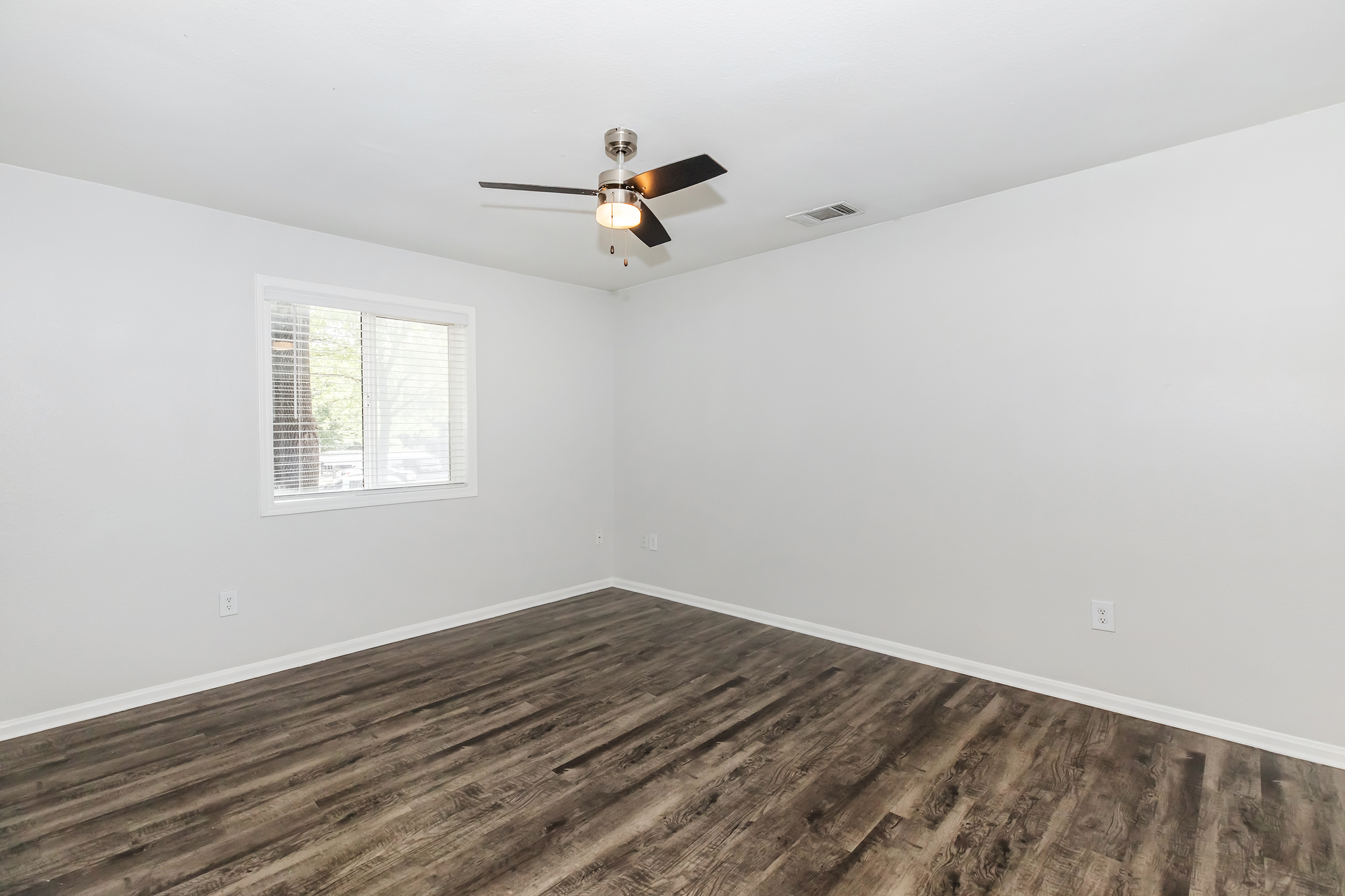 A spacious, empty room with light gray walls and dark wood-style flooring. A ceiling fan is installed, and there is a window with blinds allowing natural light to enter the space. The room is well-lit, creating an inviting atmosphere, ideal for various uses such as a living area or bedroom.