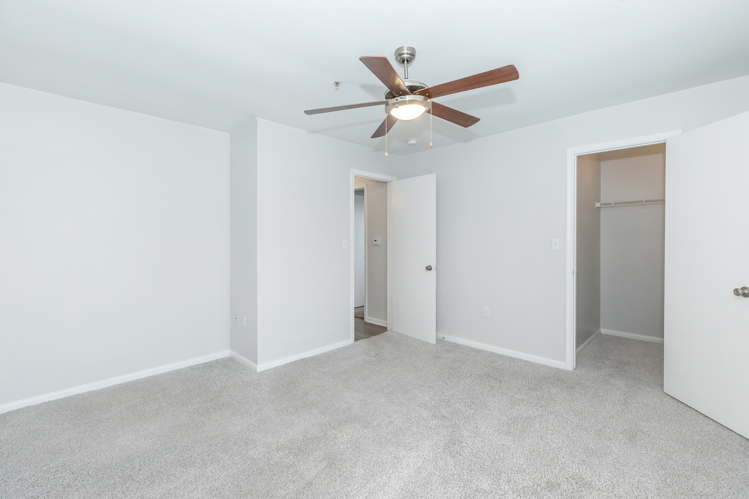 A spacious, empty room featuring light gray walls and plush gray carpeting. Ceiling fan with wooden blades is mounted above. Two open doors lead to separate areas, one leading to a closet space. Natural light brightens the room, giving it a clean and airy feel.
