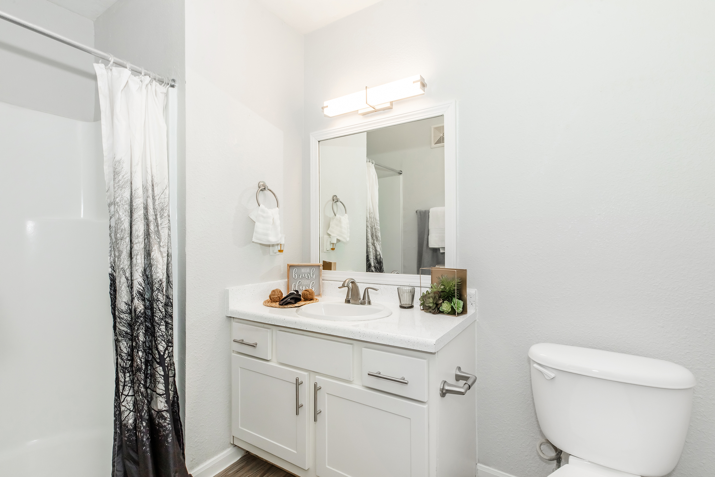A modern bathroom featuring a white vanity with a sink and storage drawers, a large mirror with overhead lighting, a white shower curtain with a tree design, a toilet, and neutral-colored walls. Decorative items include a plant and small accessories on the countertop.