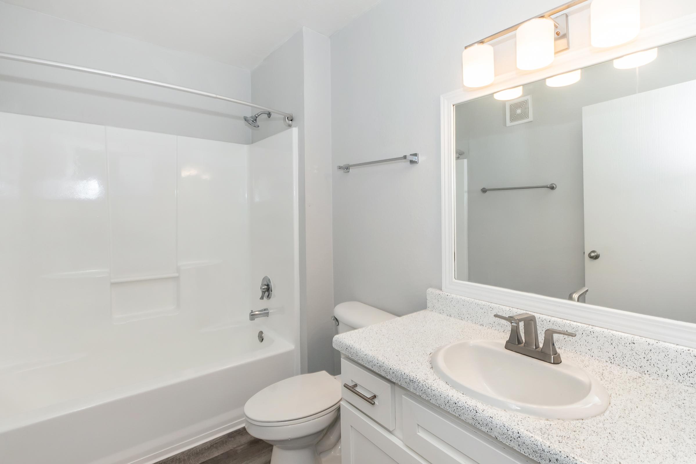 A bright, modern bathroom featuring a bathtub with a shower, white walls, and a light-colored countertop. A round mirror is above the sink, and a light fixture with three bulbs is mounted above it. The space includes a toilet and towel bar, with natural light illuminating the room.