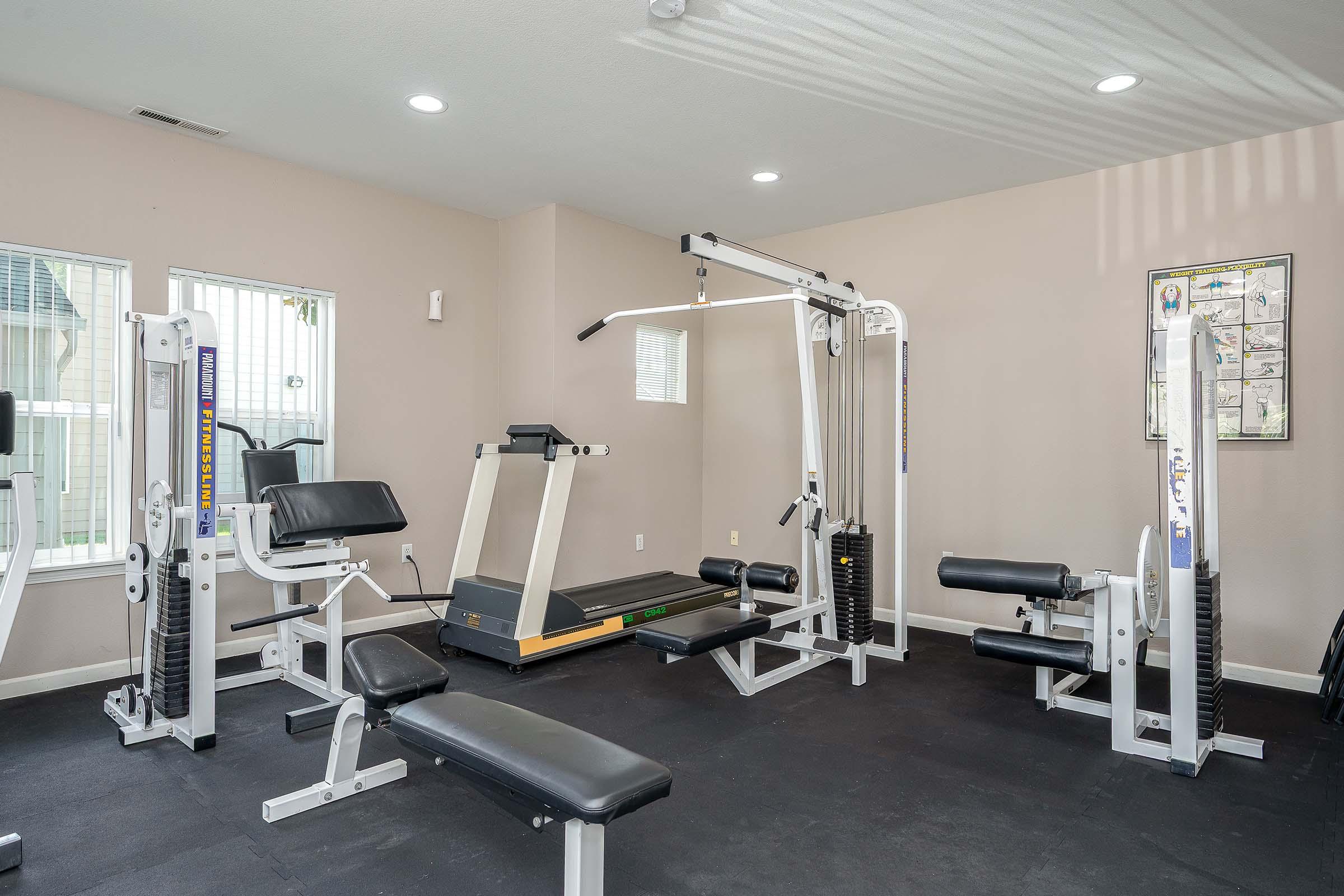 A bright and well-equipped gym featuring a multi-function weight machine, a treadmill, and various workout benches. Large windows allow natural light, creating a spacious and inviting atmosphere for exercise. The flooring is dark, providing a nice contrast to the light walls.