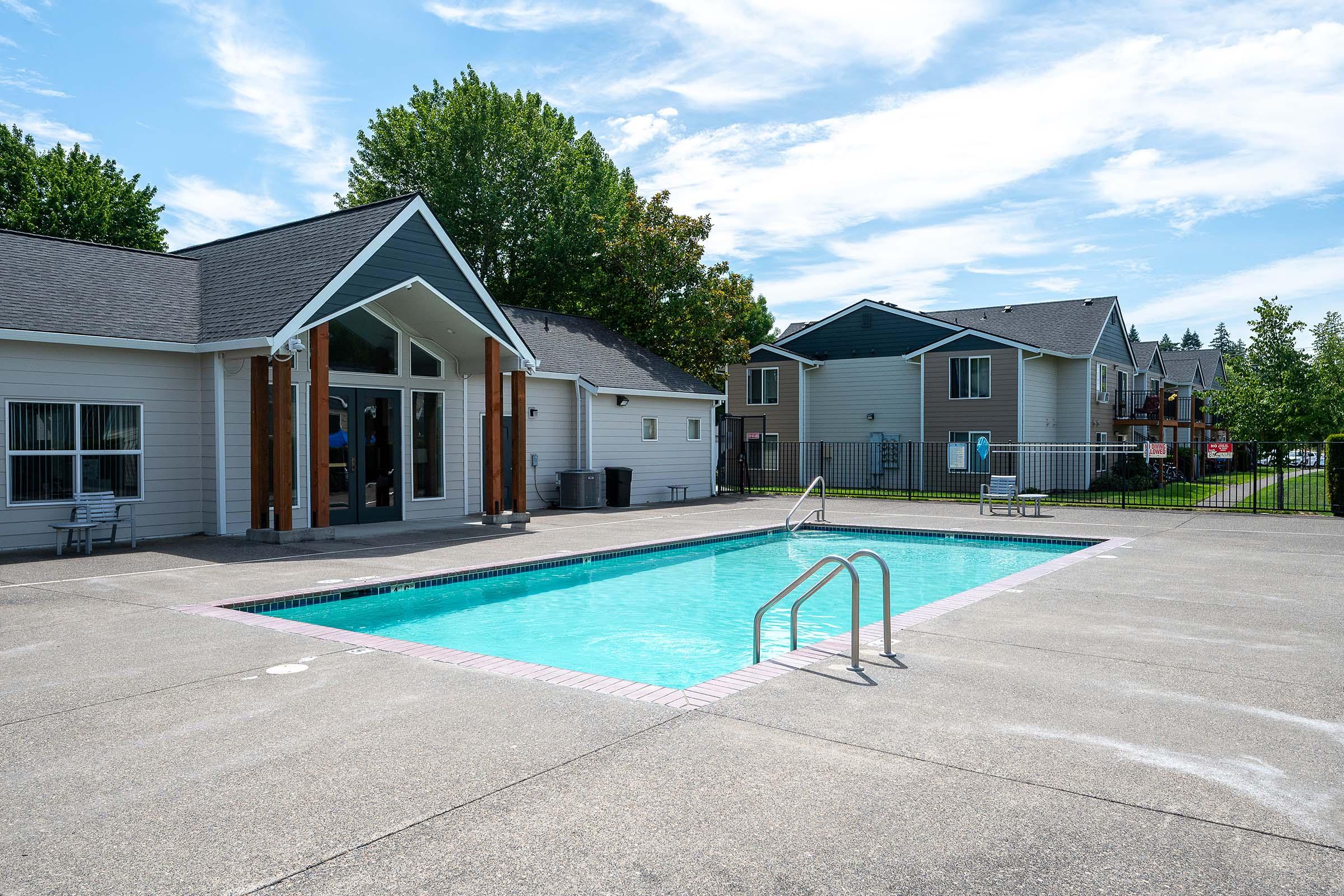 A tranquil swimming pool surrounded by a concrete patio, with a few lounge chairs nearby. In the background, there are residential buildings with green trees and a clear blue sky, creating a serene atmosphere. The pool features ladders on either side for easy access.