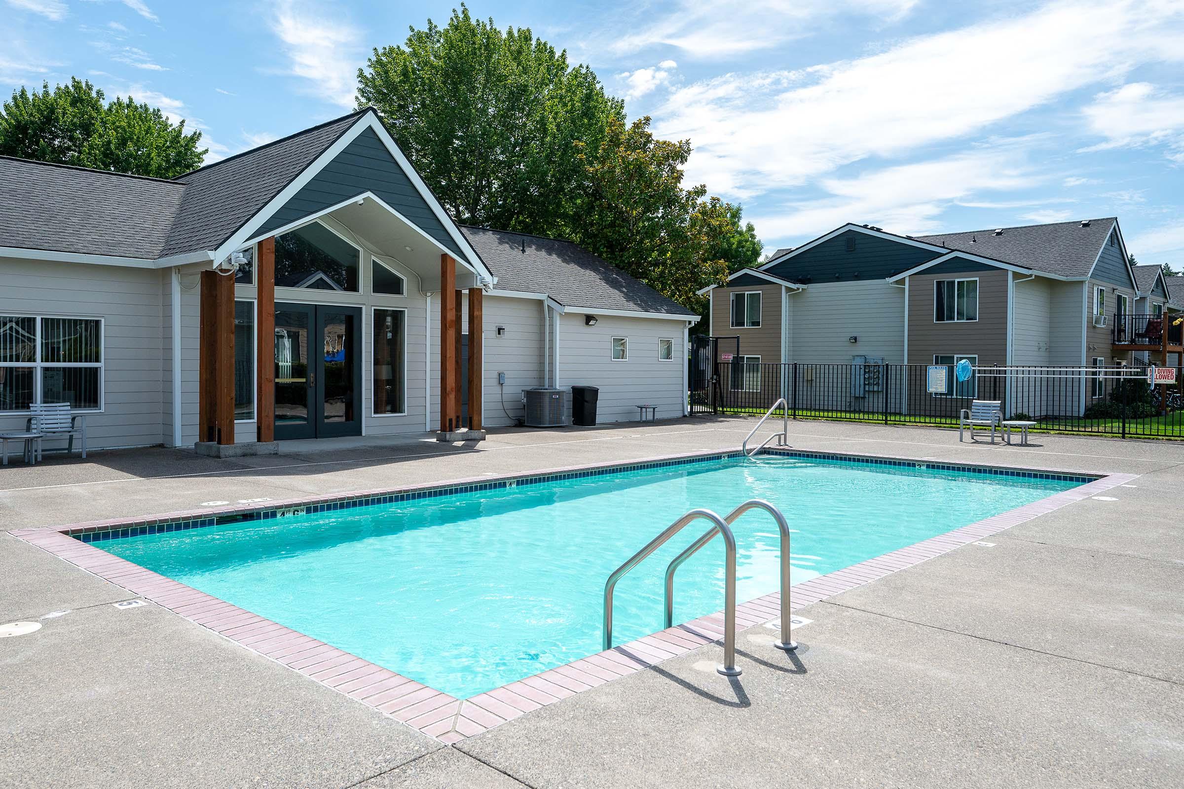 A clear swimming pool with a ladder, surrounded by a concrete patio. In the background, there are two buildings with multiple apartments, and a fence enclosing the area. Lush green trees provide shade, and the sky is partly cloudy.