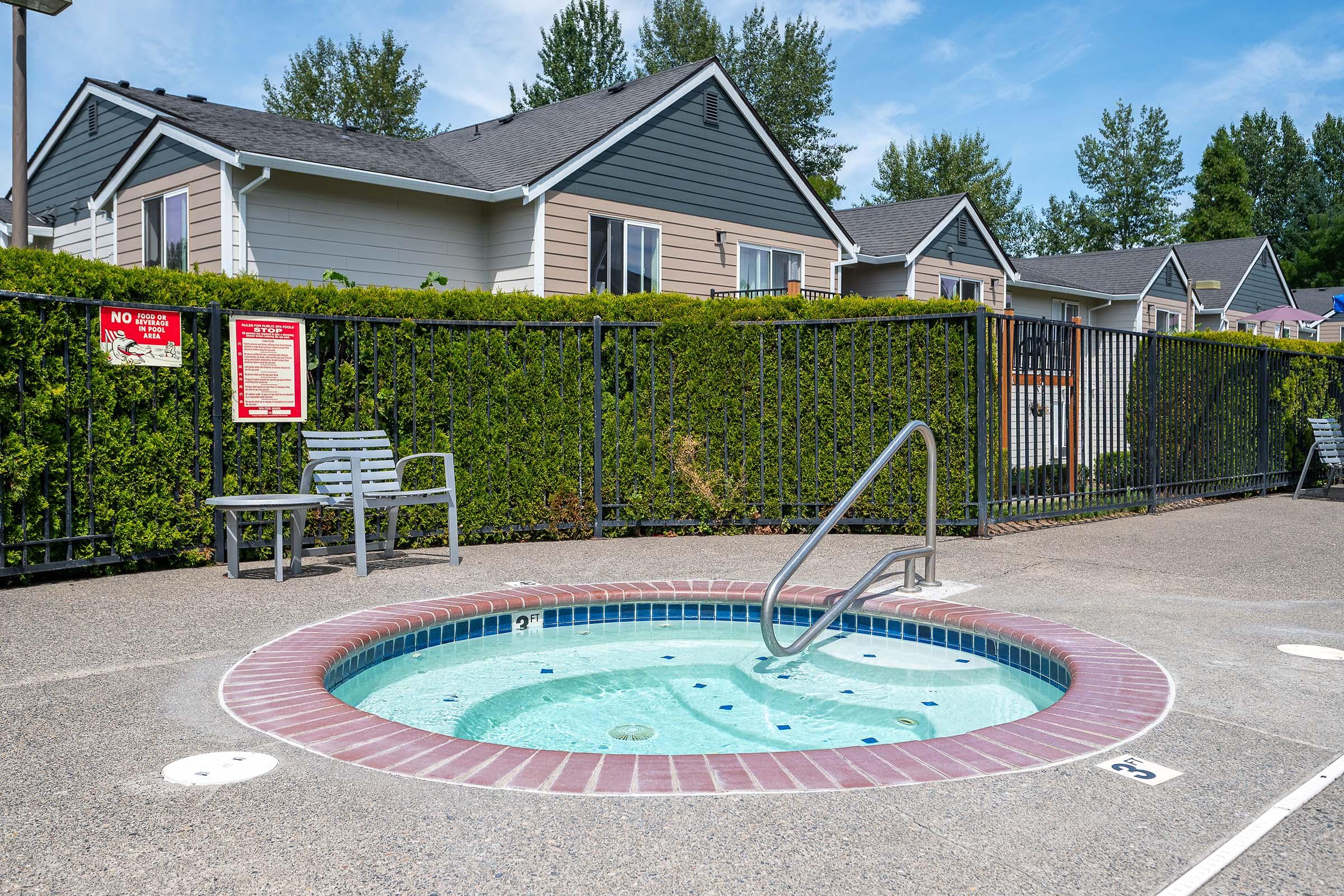A small outdoor hot tub with a metal handrail, surrounded by a low fence and a neatly trimmed hedge. Nearby, there are two metal chairs and a sign that reads "NO DIVING." In the background, several residential buildings can be seen against a clear blue sky.