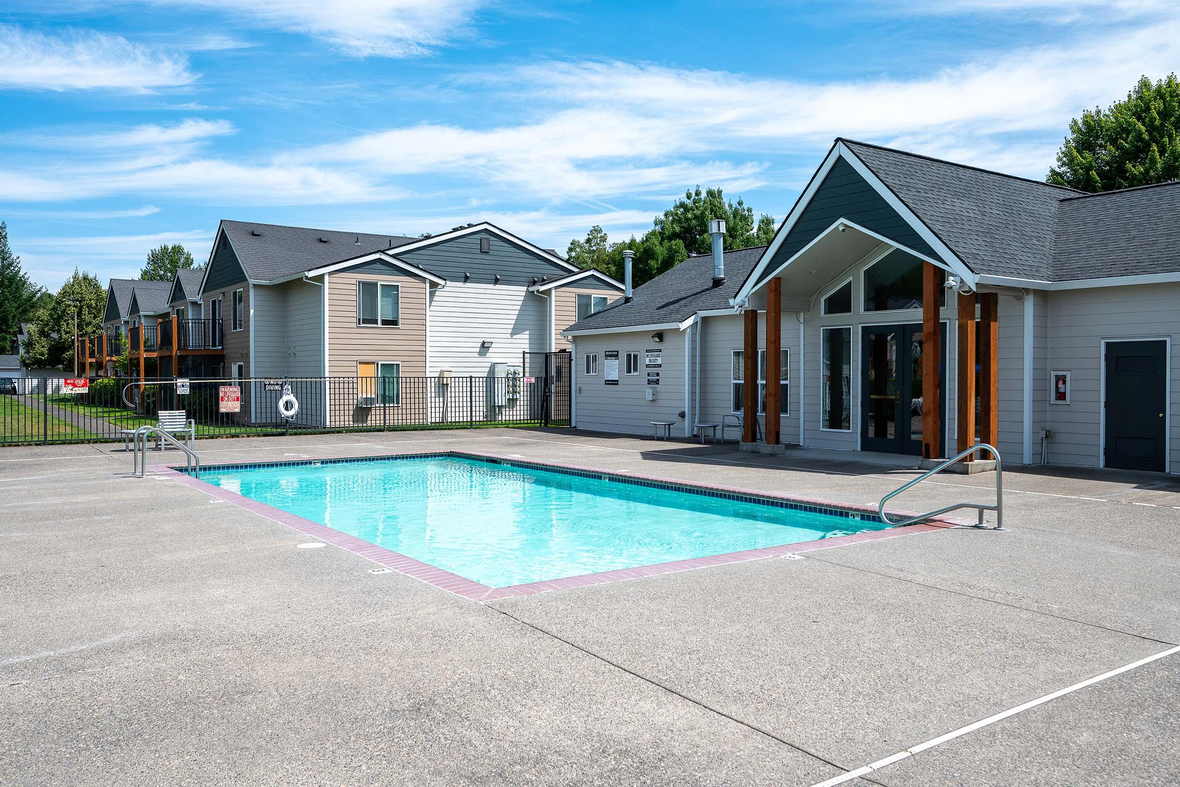 A clear blue swimming pool surrounded by a concrete deck, located near residential buildings. The area is sunny with a few trees in the background and a fenced perimeter. A building with large windows is adjacent to the pool, indicating a recreational or communal space.