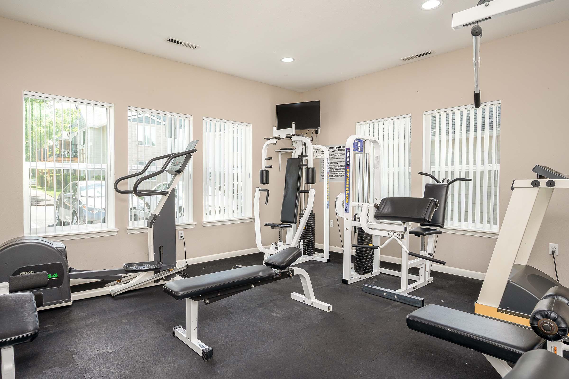 A fitness room featuring various exercise equipment, including a treadmill, multi-station weight machine, stationary bike, and a bench with weights. The room has large windows allowing natural light and a television mounted on the wall. The floor is black and the walls are light-colored.