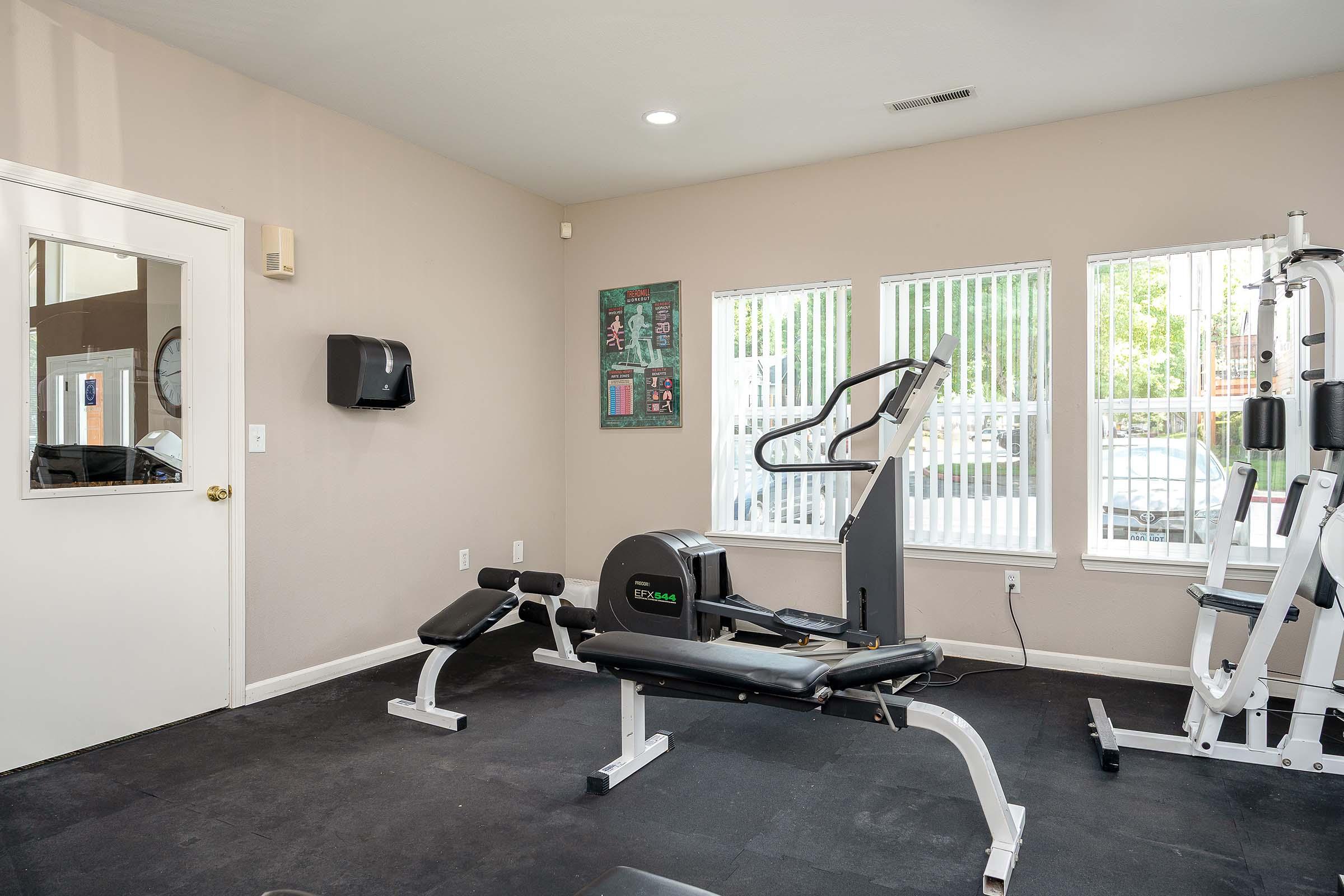A well-lit gym with exercise equipment, including a stationary bike, a weight bench, and a multi-gym machine. Large windows allow natural light to fill the space, and there is a wall-mounted mirror and a paper towel dispenser visible. The flooring is black rubber, creating a functional workout environment.