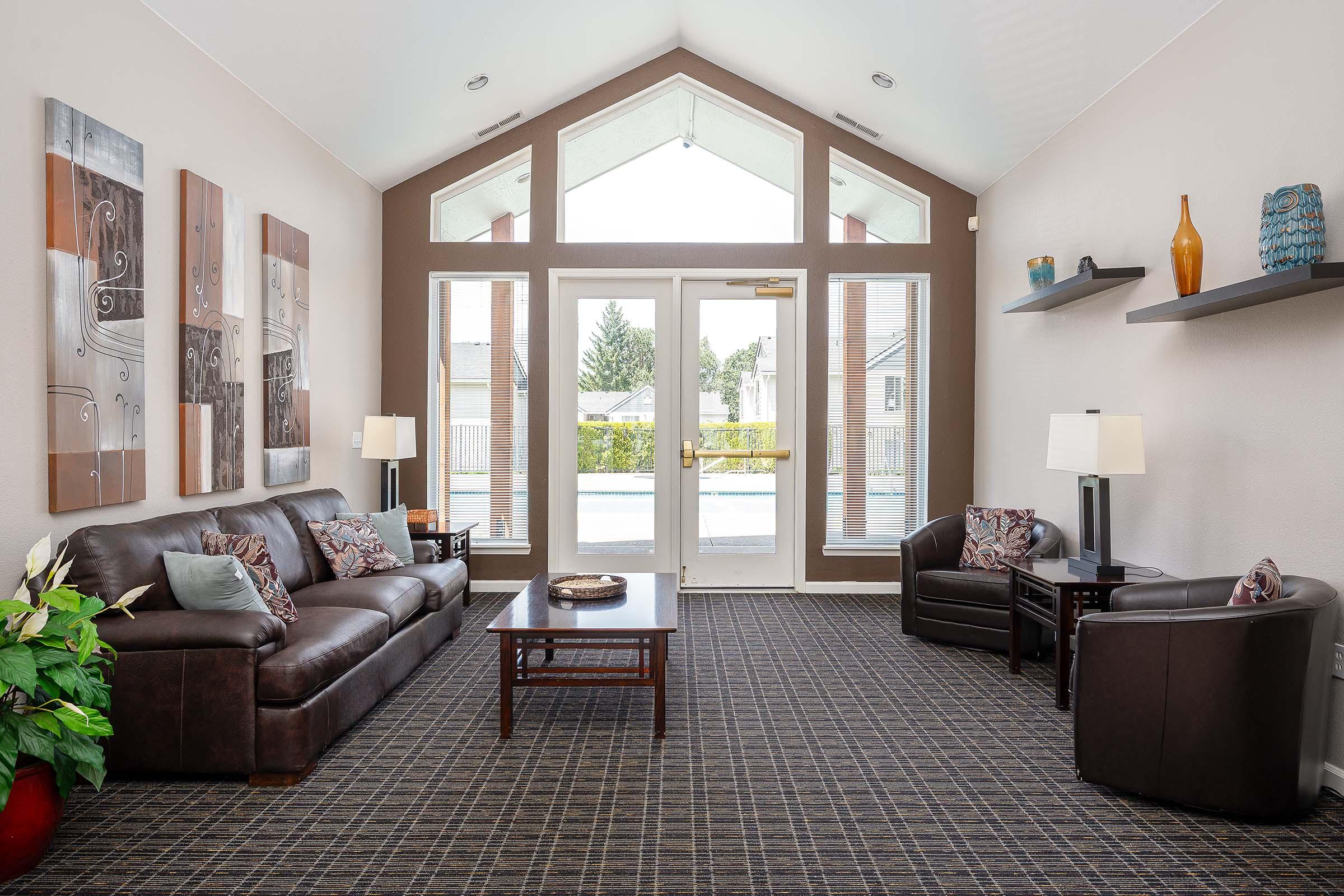 A spacious, well-lit living room featuring a brown leather sofa and armchair, with a coffee table in the center. The walls are decorated with abstract art, and there are decorative shelves with vases. Large windows provide a view of the outdoors, enhancing the inviting atmosphere.