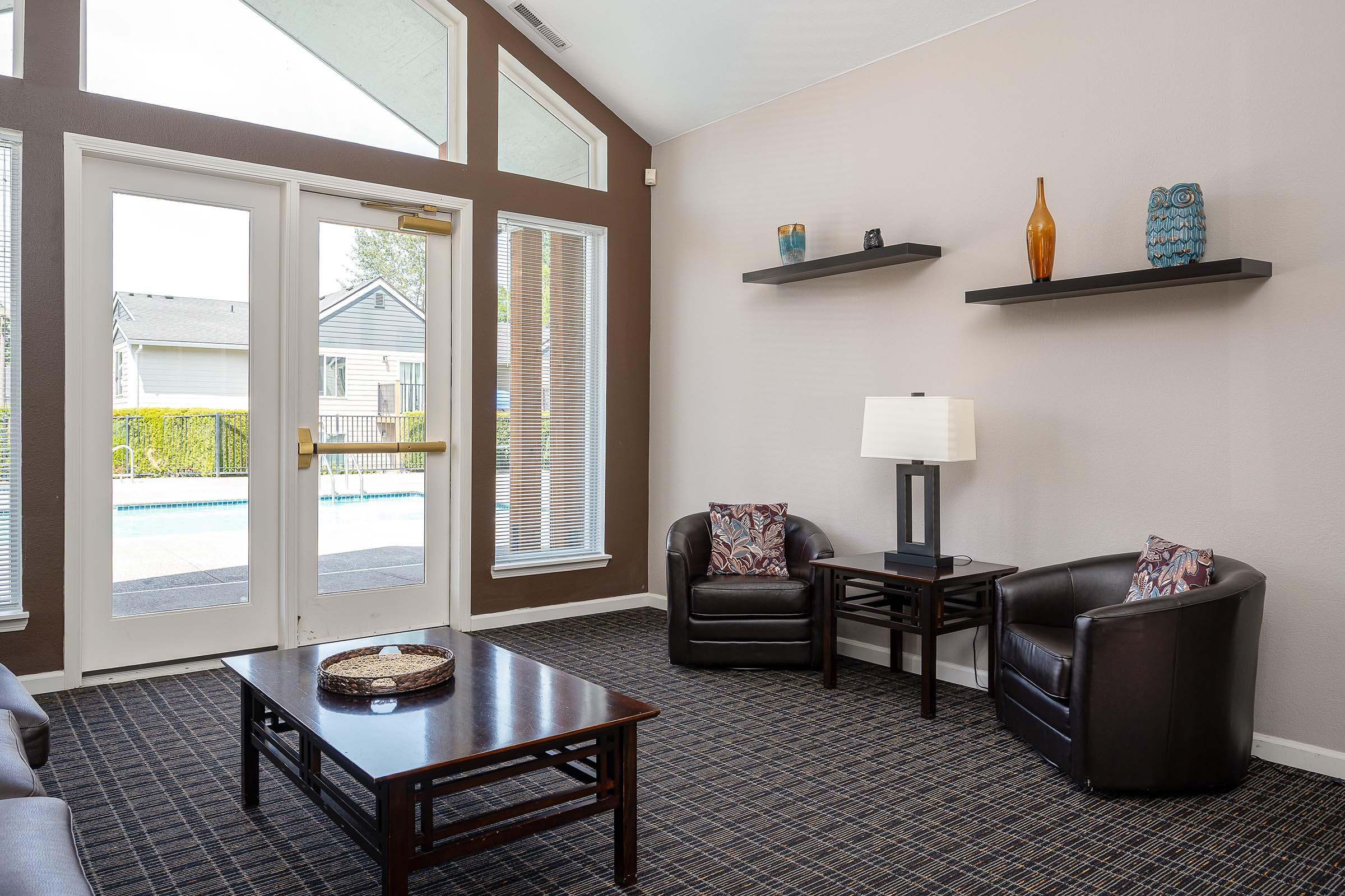 A cozy indoor seating area featuring two brown leather armchairs, a wooden coffee table, and a lamp on a side table. Shelves adorned with decorative vases are mounted on the wall, and large windows provide a view of an outdoor pool area. The carpet has a patterned design, enhancing the inviting atmosphere.