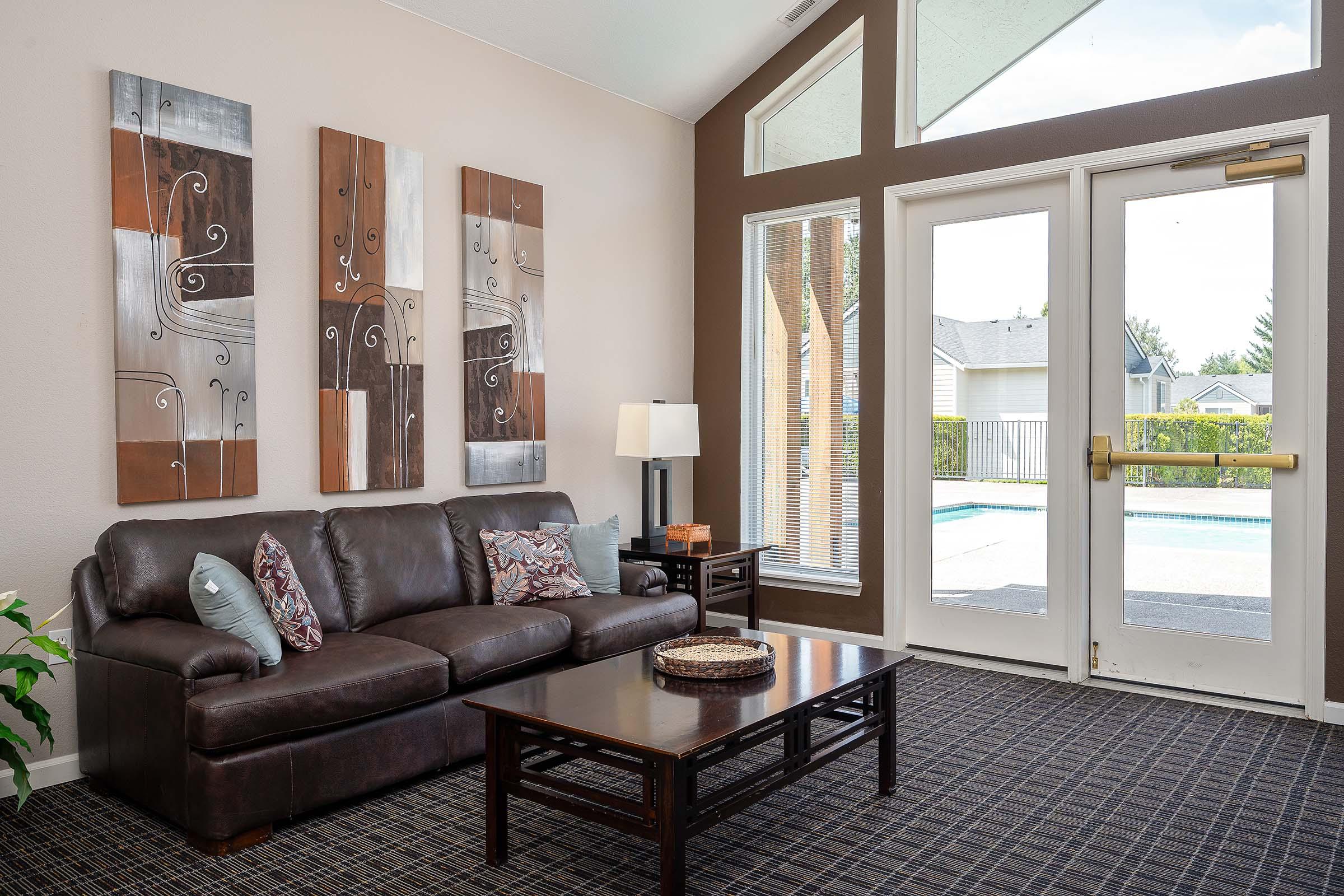 A cozy living room featuring a dark leather sofa with decorative pillows, a wooden coffee table, and artwork on the walls. Large windows let in natural light, showcasing a view of an outdoor pool area. The flooring is carpeted in a patterned design, adding warmth to the space.