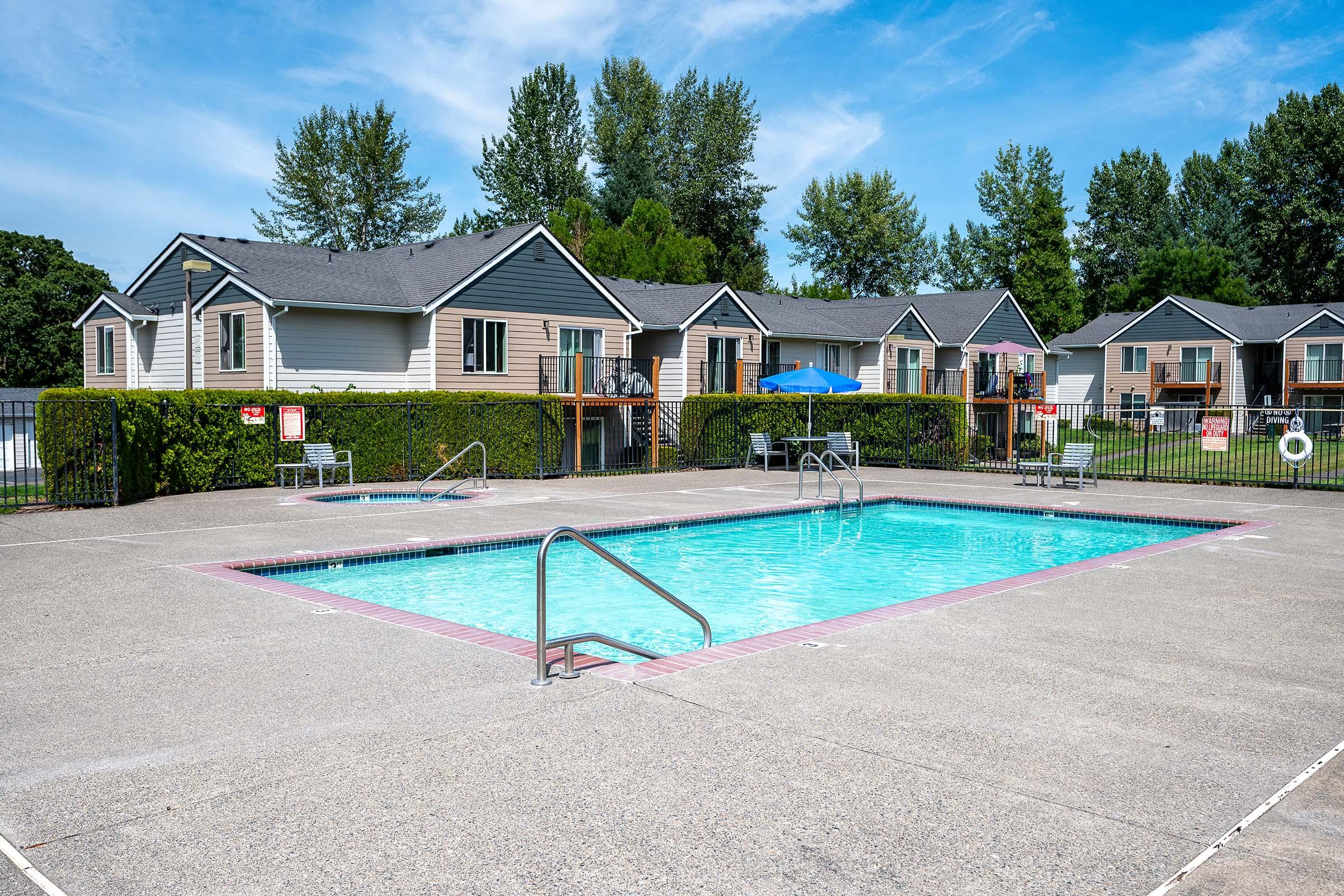 A clear swimming pool surrounded by a concrete deck, with a ladder on one side. In the background, there are two-story residential buildings with a mix of green lawns and hedges. A blue umbrella provides shade near the pool, and there are signs nearby, indicating pool rules. Bright, sunny sky above.