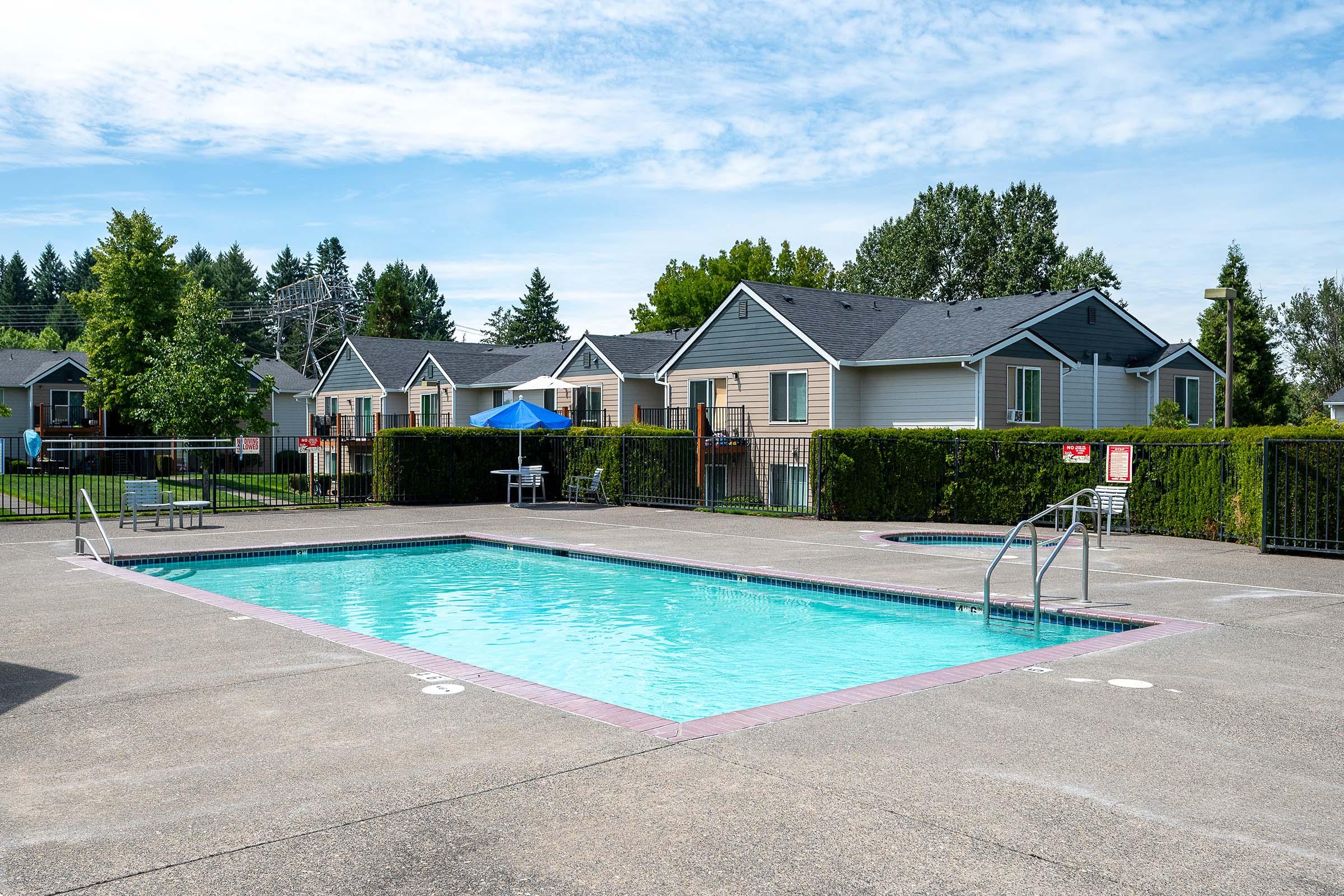 A clear, inviting outdoor swimming pool surrounded by a concrete patio. There are lounge chairs positioned near the pool, and a shaded area with an umbrella. In the background, several residential buildings are visible, along with green trees and shrubs lining the property. The sky is bright with a few scattered clouds.