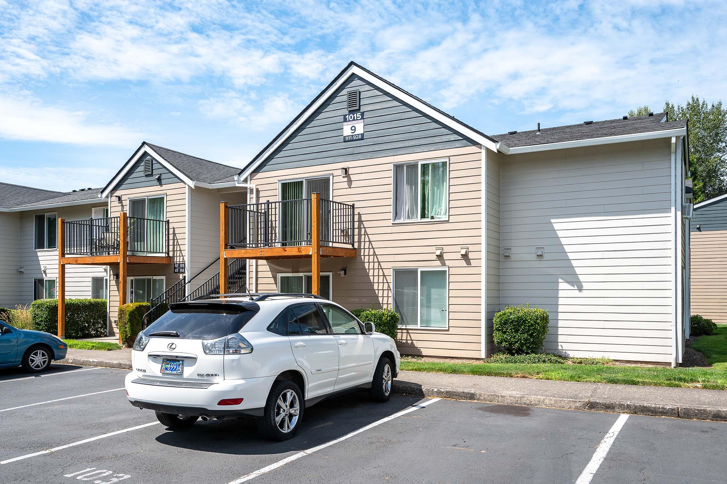 A two-story apartment building with a light exterior and dark gray accents. The building features two balconies, green curtains visible through the windows, and several parking spaces in front. A white SUV is parked in the foreground, with another blue vehicle further down the lot. The sky is partly cloudy.