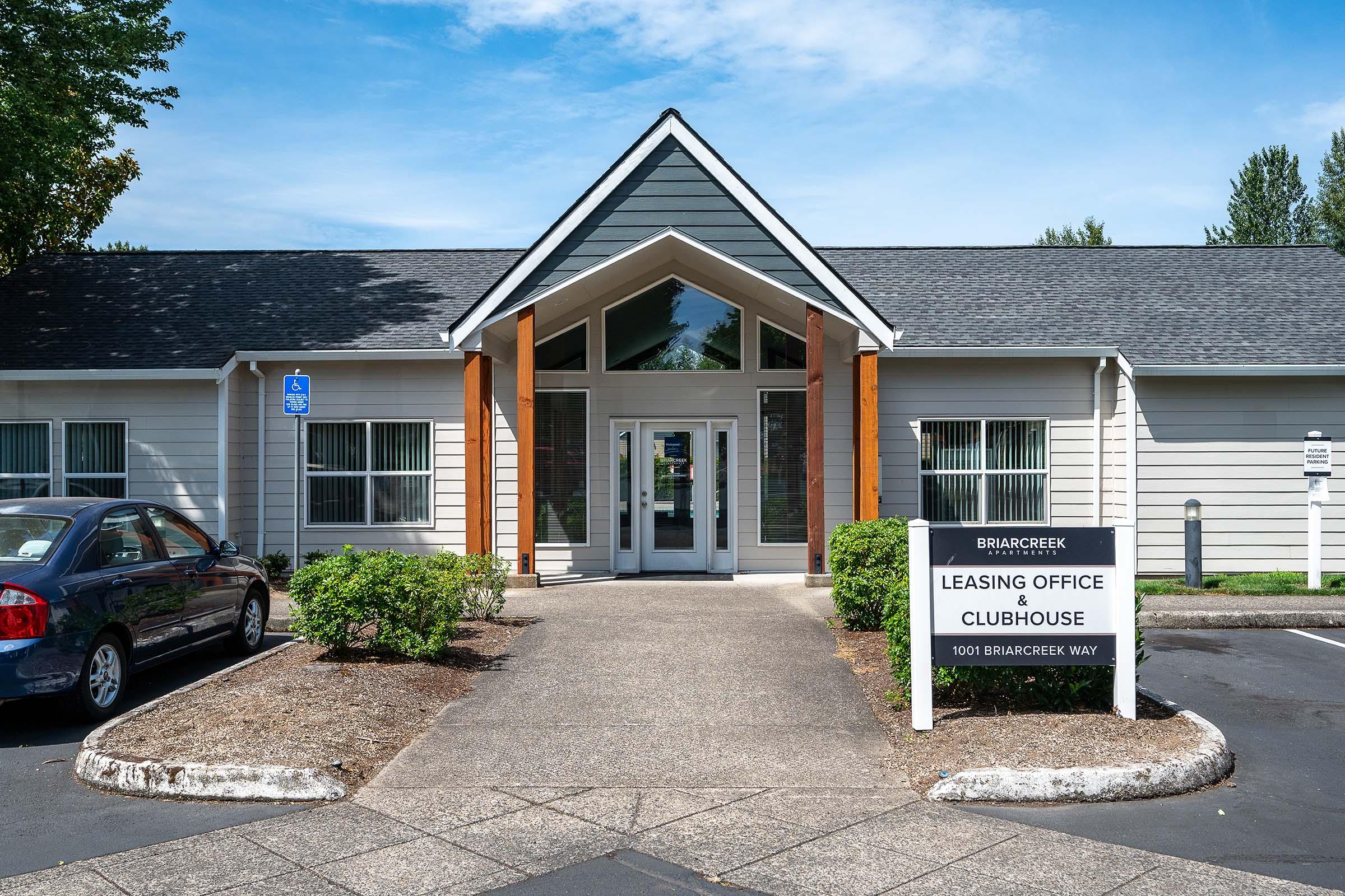A modern leasing office building with a peaked roof, large front windows, and a sign that reads "Leasing Office" and "Clubhouse." The exterior features a mix of gray and wooden accents, surrounded by landscaped shrubs and trees. A parking lot is visible in front, with a car parked nearby.