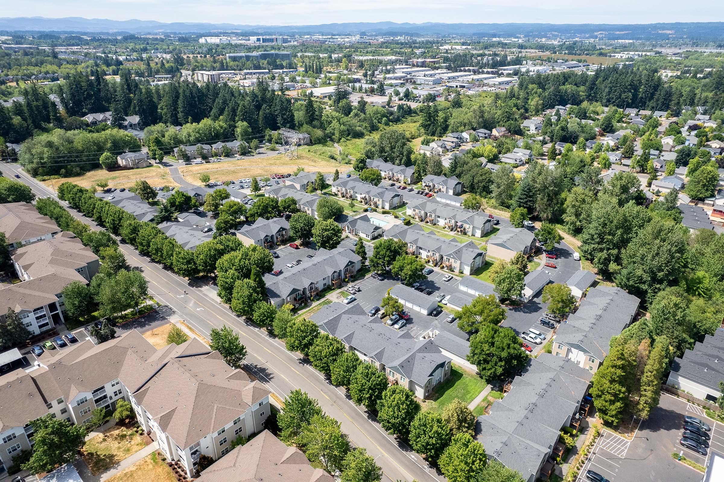 Aerial view of a suburban neighborhood featuring rows of residential buildings with dark rooftops, surrounded by trees and green spaces. A road runs along the edge of the area, with parking lots visible. In the background, there are more residential and commercial developments, under a clear blue sky.
