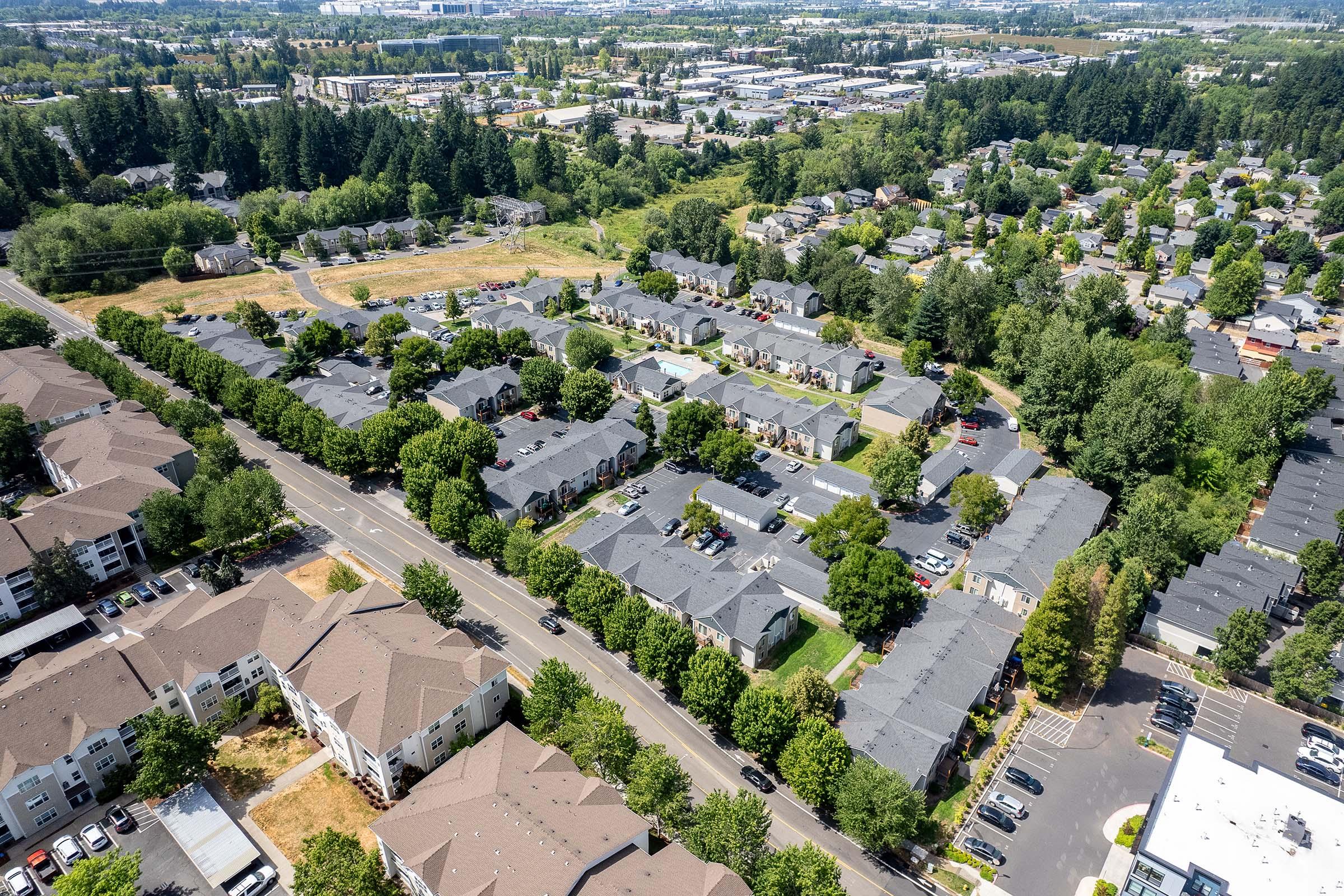 Aerial view of a suburban neighborhood featuring rows of residential buildings surrounded by green trees and landscaped areas. Roads and parking lots are visible, with some vehicles parked along the streets. In the background, industrial and commercial buildings can be seen.