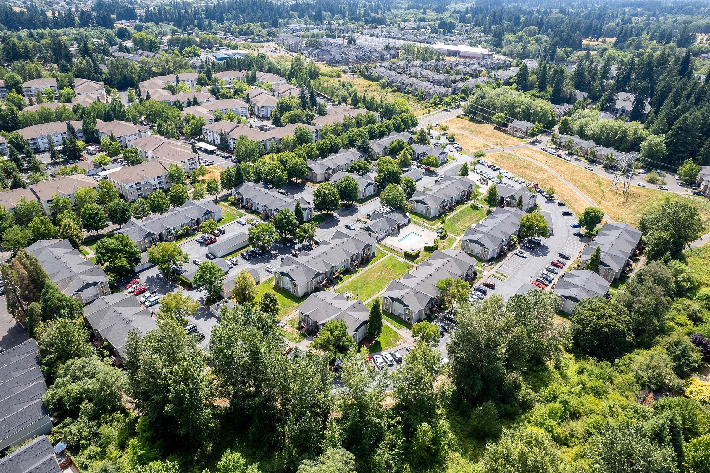 Aerial view of a residential neighborhood featuring multiple apartment complexes, green trees, and organized parking lots. The landscape includes grassy areas and trails, with additional housing developments visible in the background. A clear blue sky complements the scene.
