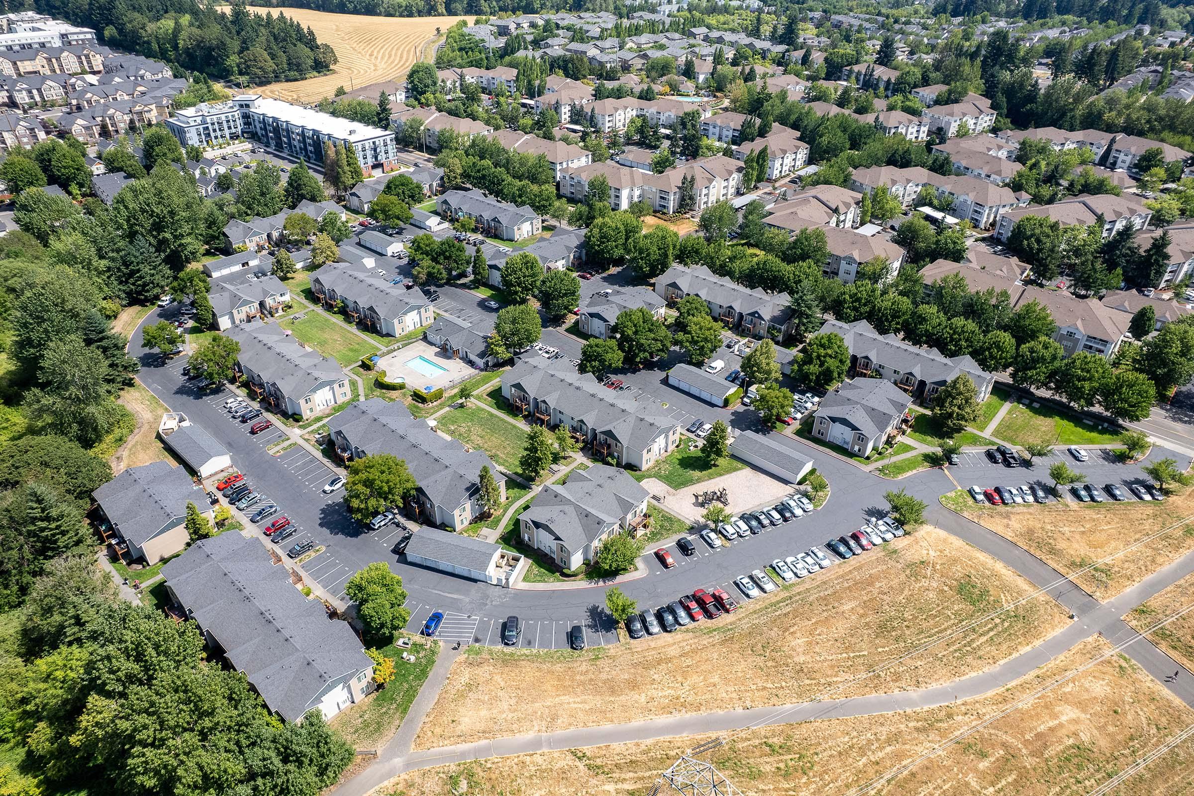An aerial view of a residential neighborhood featuring clusters of apartment buildings, green spaces, and a swimming pool. Surrounding roads with parked cars are visible, alongside patches of greenery and agricultural land in the distance. The layout suggests a well-planned community with close-knit living spaces.