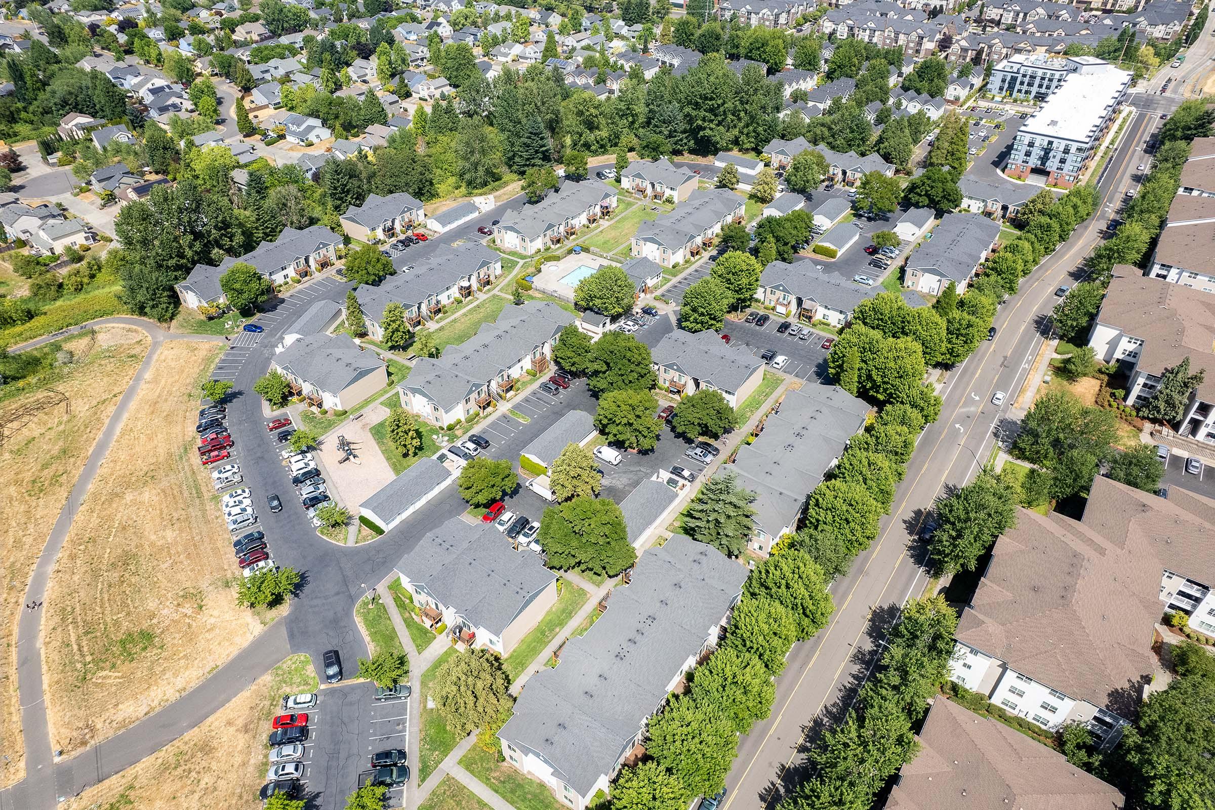 Aerial view of a residential neighborhood featuring multiple apartment buildings surrounded by greenery and parking areas. The layout includes winding streets, trees, and parked cars, with a larger building visible in the top right corner.
