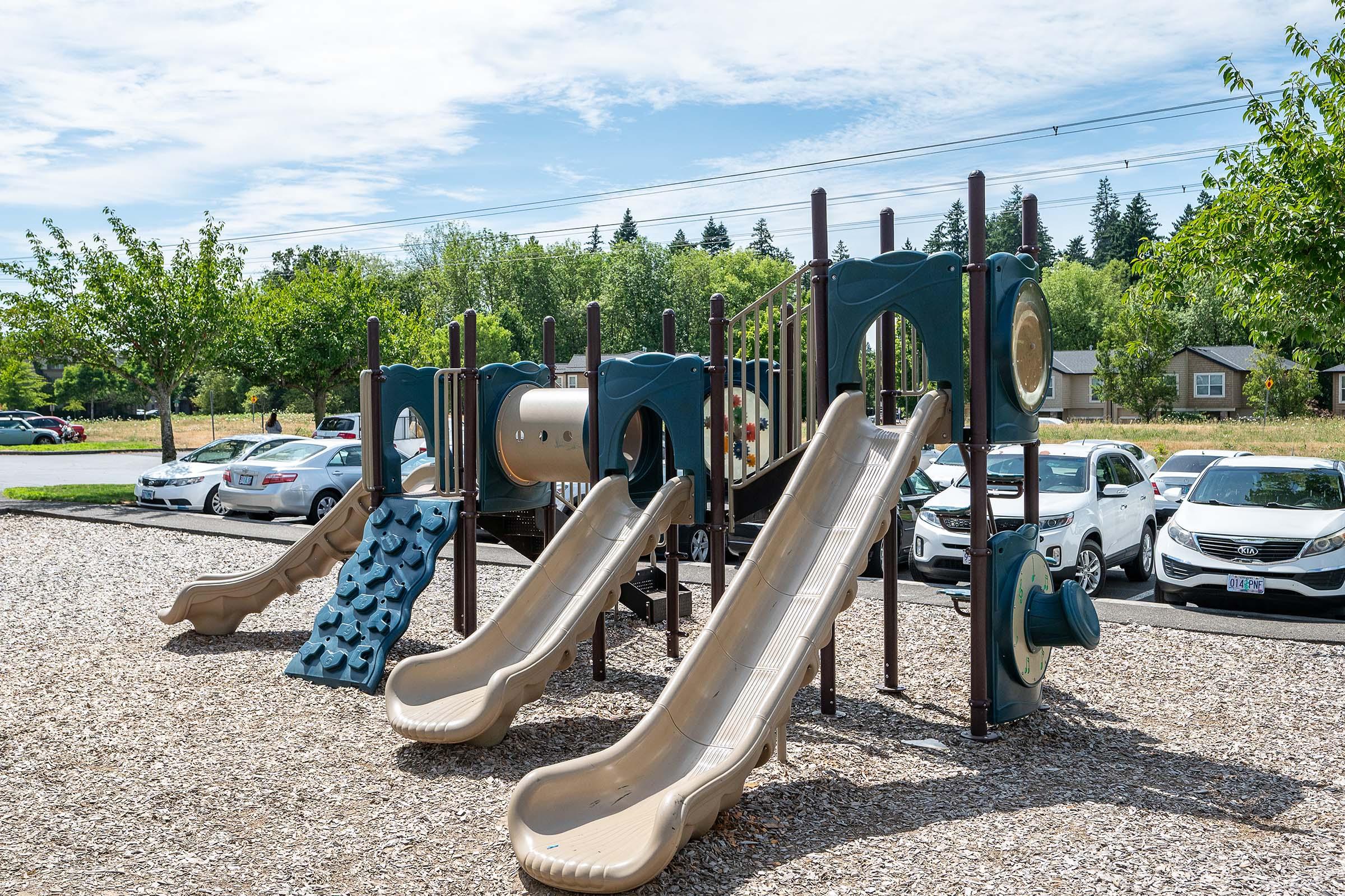 Playground equipment featuring two slides, a climbing wall, and various play structures situated on a bed of wood chips. In the background, several cars are parked, with greenery and trees visible nearby under a partly cloudy sky.