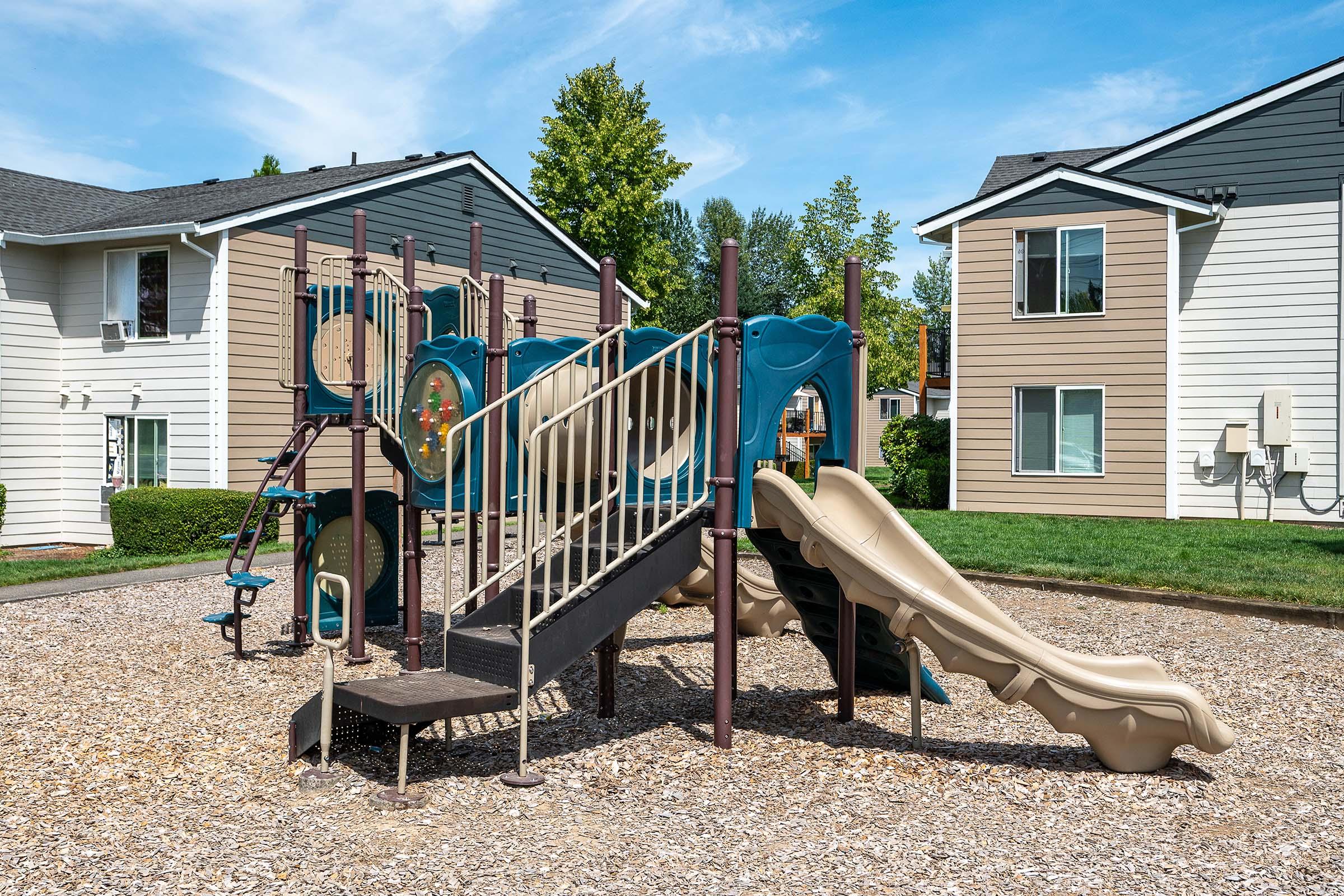 A colorful playground with climbing structures and slides, set on a gravel surface. In the background, two residential buildings with multiple windows are visible, surrounded by greenery and blue sky. The scene is bright and inviting, ideal for children to play.