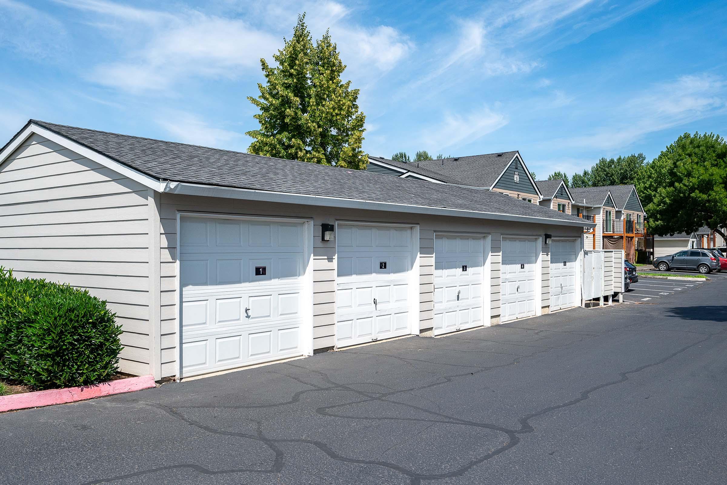 A row of four white garage doors attached to a light-colored building. The garages are set against a backdrop of a blue sky with a few clouds and green trees in the distance. A parking lot is visible in front of the garages, with a neatly paved blacktop surface.