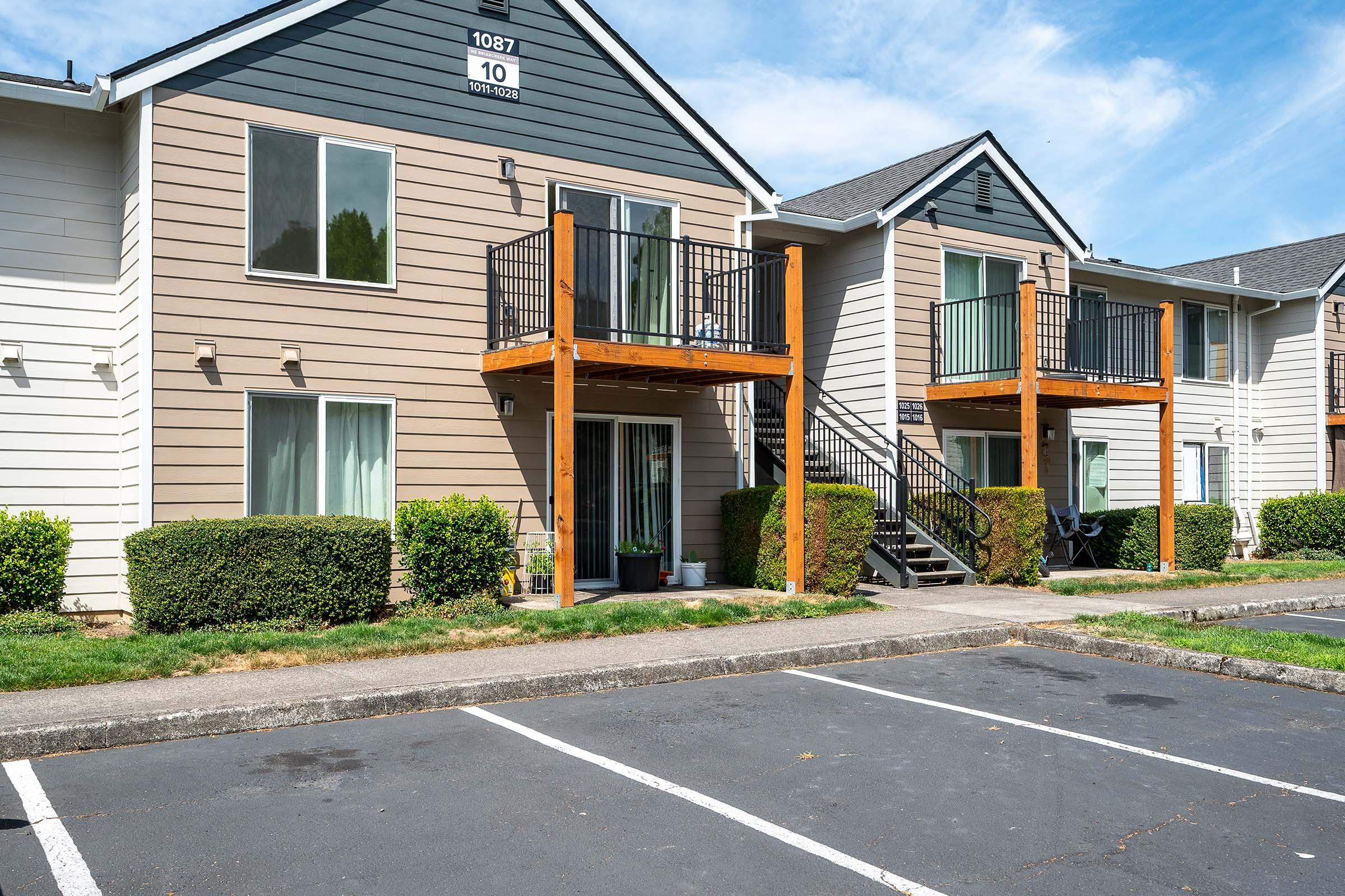 Two multi-story apartment buildings with balconies. The building on the left has a tan exterior with dark trim, while the building on the right features a light-colored exterior. The area includes a parking lot with empty spaces, manicured shrubs, and a clear blue sky overhead.
