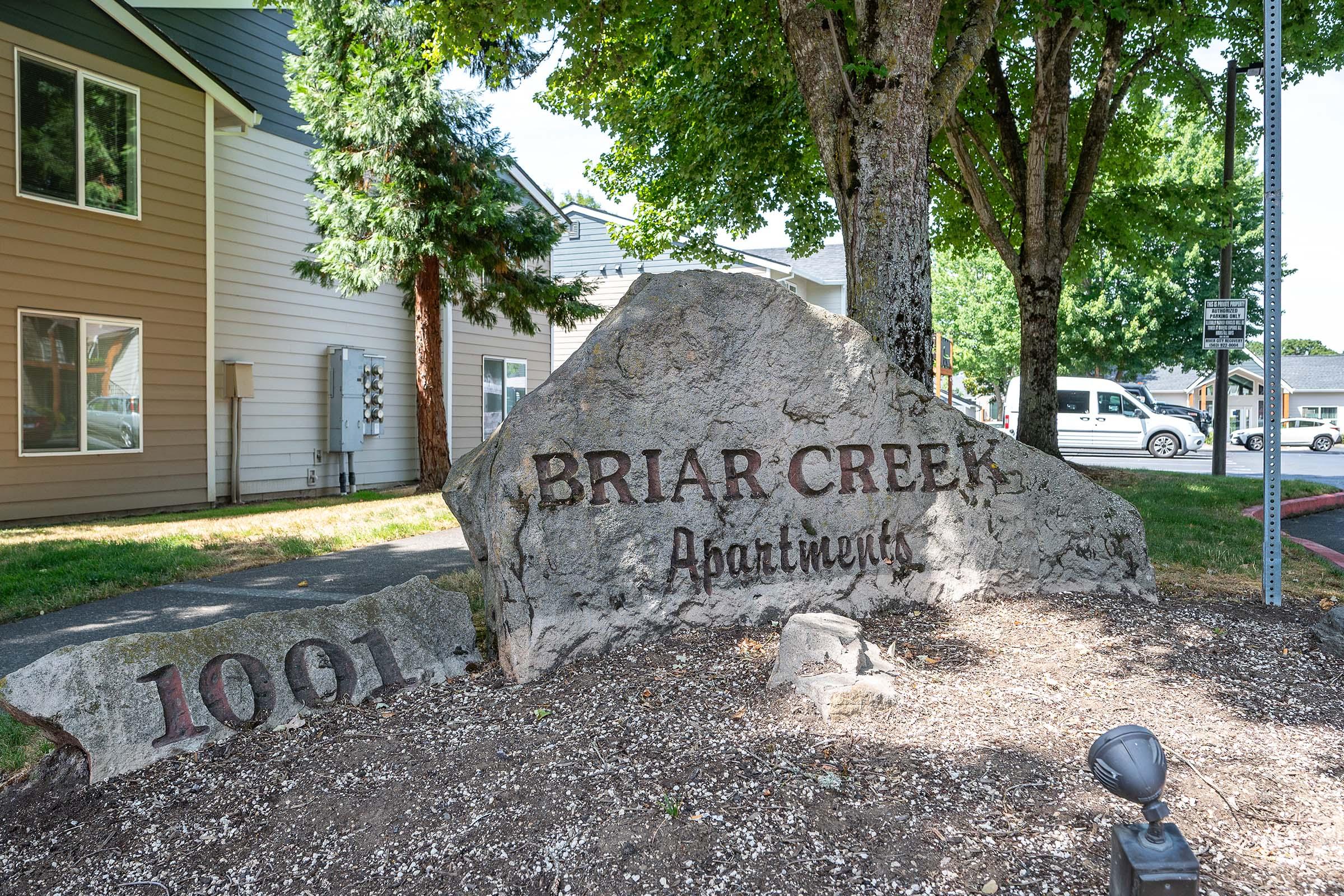 Sign on a large rock displaying "Briar Creek Apartments" and the number "1001," surrounded by greenery and apartment buildings in the background.