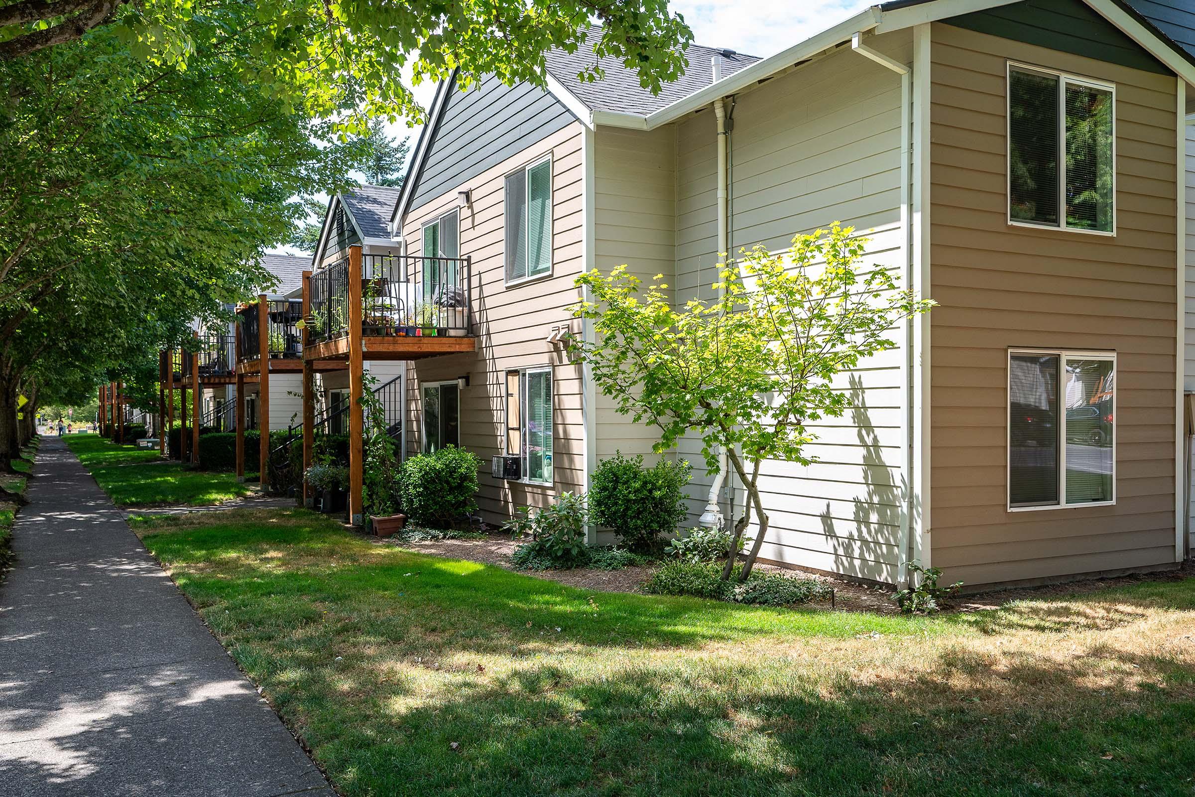 Two-story residential building with a light-colored exterior and green landscaping. There are balconies on the first and second floors, surrounded by trees. A paved walkway runs alongside the building, leading through a grassy area. Sunlight is bright, creating a warm atmosphere.