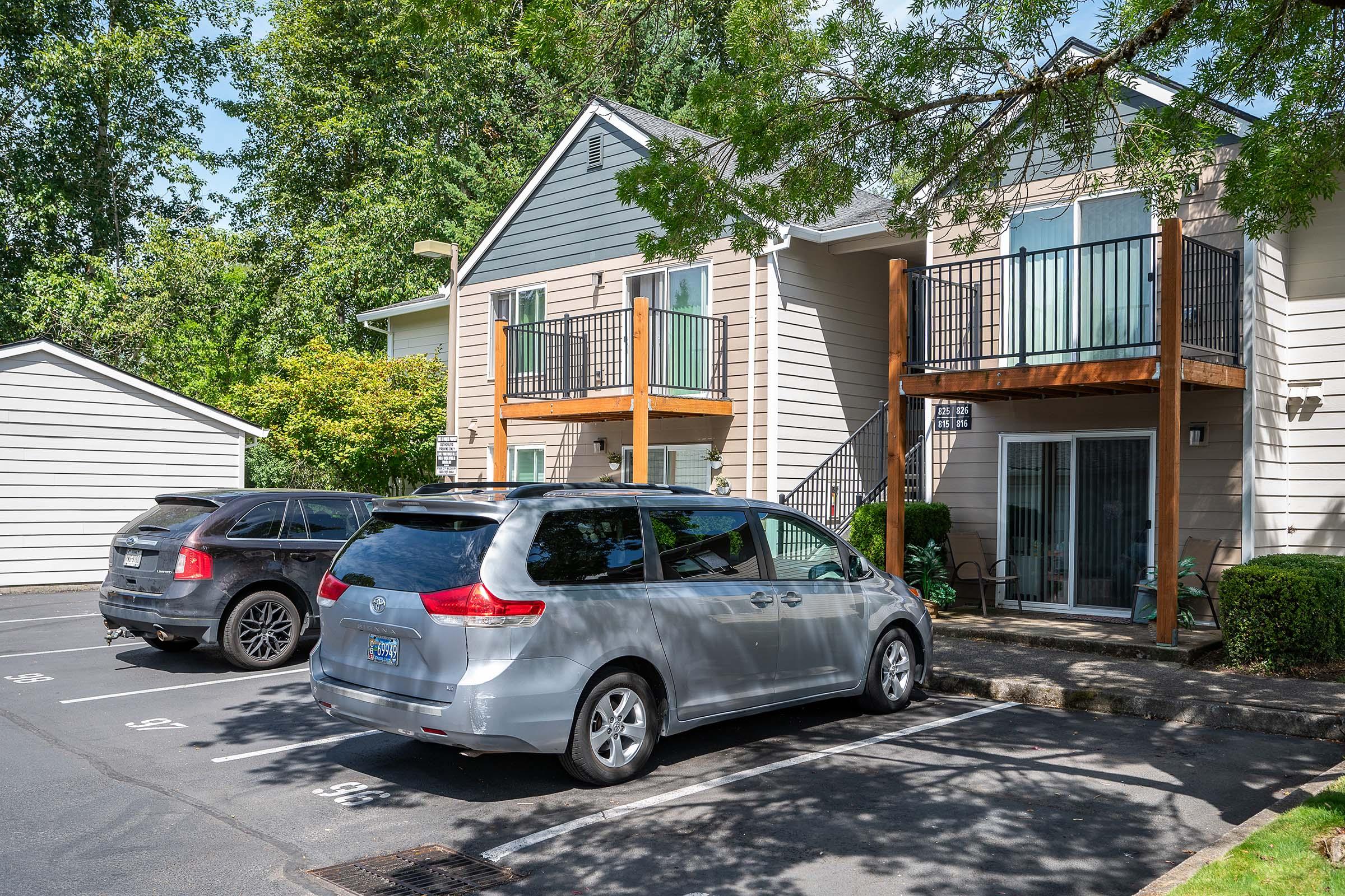 Two parked cars in the foreground—one black SUV and one silver minivan—visible in a parking lot. In the background, a two-story apartment building features a light gray exterior with wooden balconies and a small porch. Lush green trees and bushes surround the area, creating a pleasant residential atmosphere.