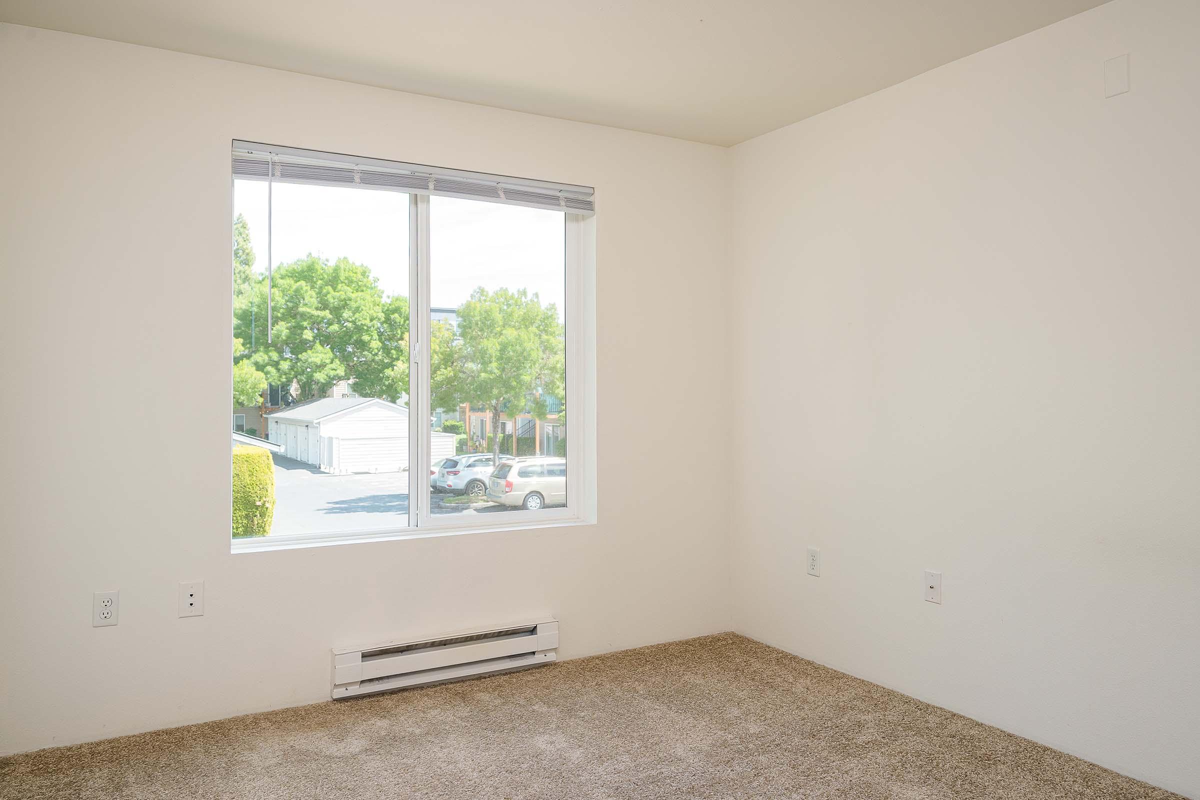 A bright, empty room with white walls and a large window. The window offers a view of green trees and parked cars outside. The floor is covered with light brown carpet, and there are no visible furnishings or decorations. Heating vents are present on the walls.