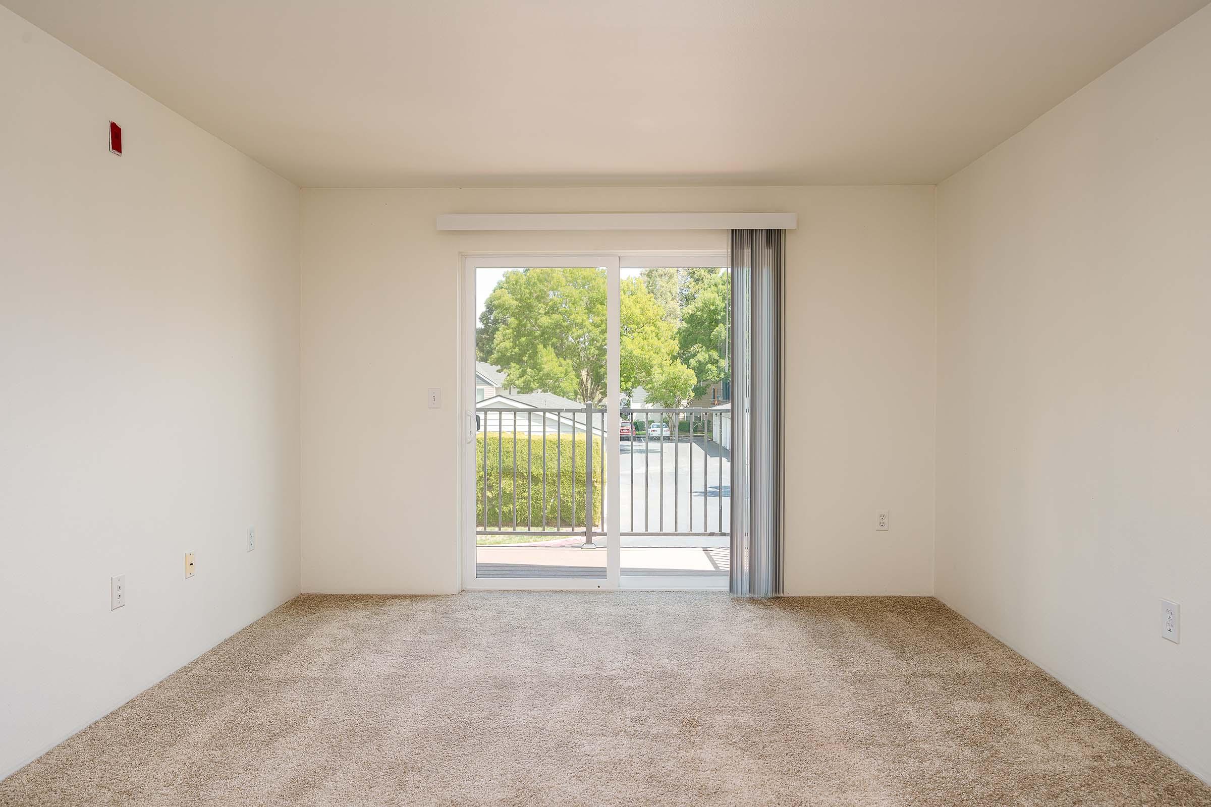 Empty room with beige carpet and white walls, featuring a sliding glass door that opens to a balcony or patio area. Natural light streams in through the door, illuminating the space. There are no furnishings or decorations present, creating a clean and minimalistic appearance.