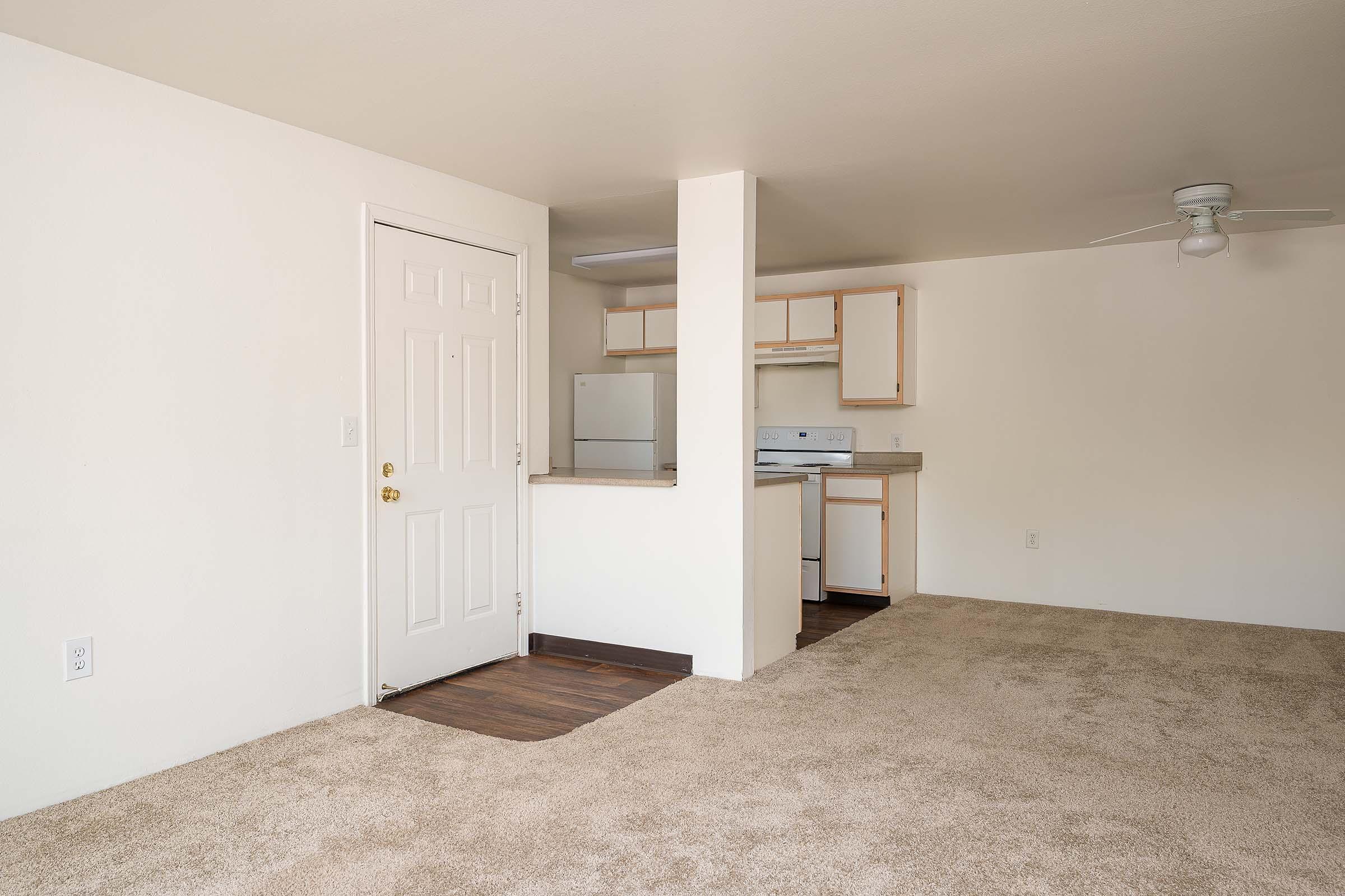 A spacious, empty living area with beige carpet and white walls. The entrance door is visible on the left, and a small kitchen area is in the background, featuring wooden cabinets and appliances. A ceiling fan hangs above, and natural light illuminates the room.