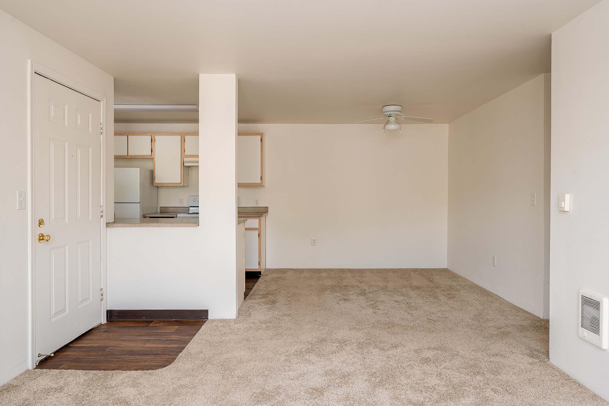 A spacious, empty living room area featuring beige carpet and white walls. There is a ceiling fan, and a doorway leads to a kitchen with wooden cabinets in the background. Natural light filters in, creating a bright and inviting atmosphere.