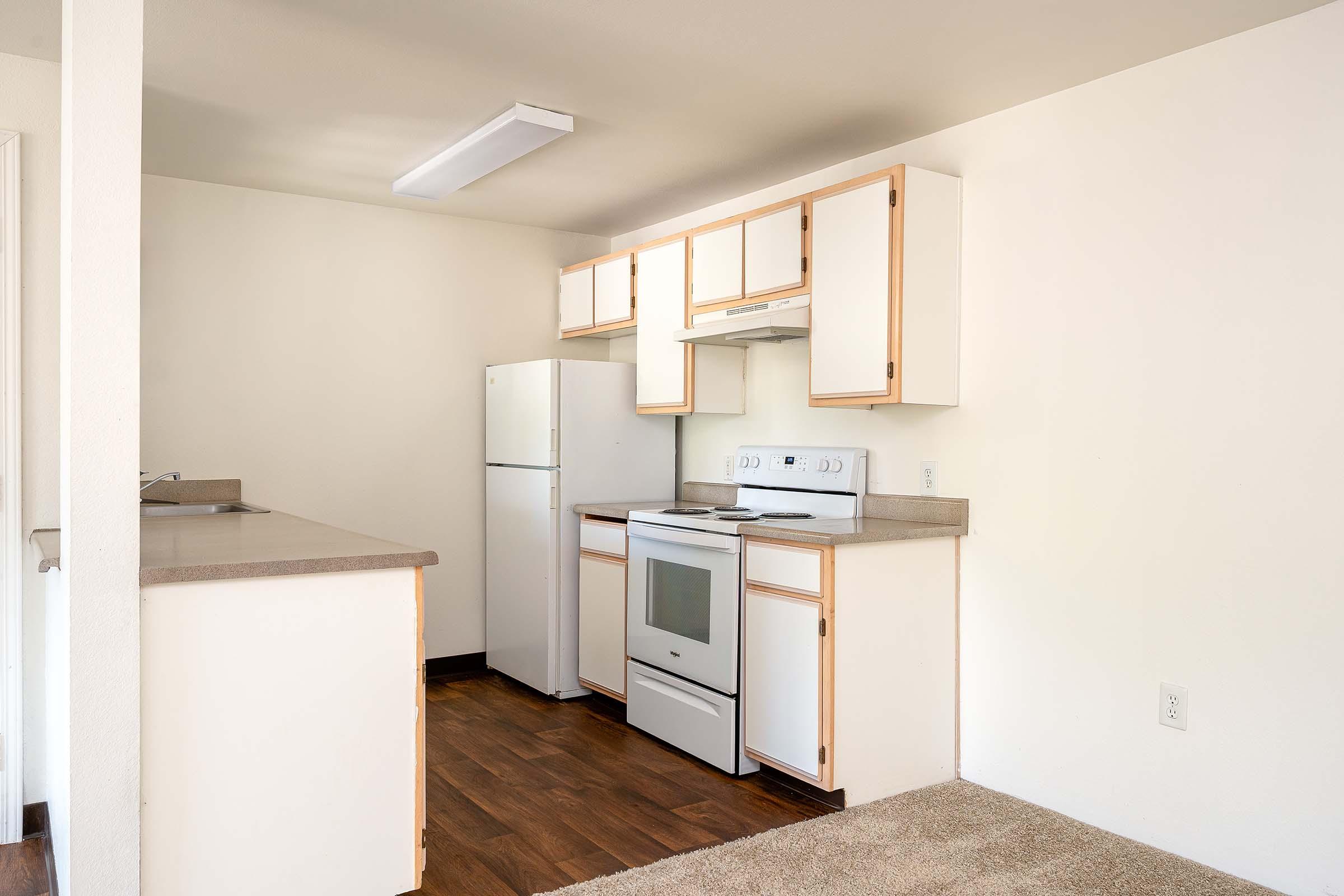 A modern kitchen featuring white cabinetry, a white stove and refrigerator, and a light countertop. The floor is wooden, and the walls are painted a light color. A small countertop extends from the kitchen area, providing additional workspace. The overall design is clean and minimalist.