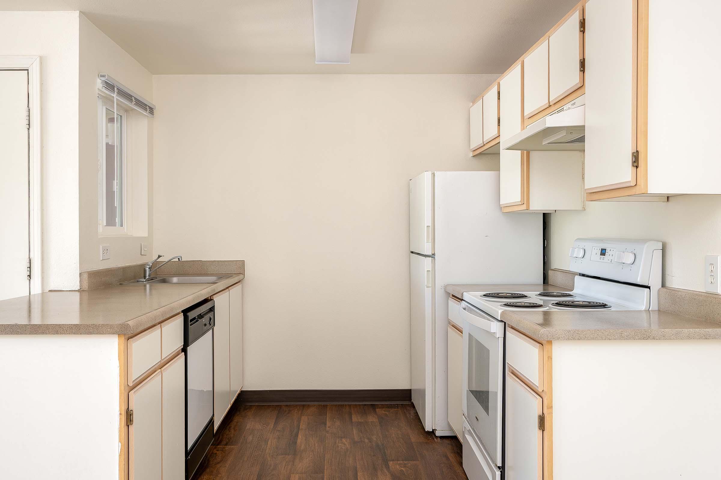 A bright and clean kitchen featuring white cabinetry, a dishwasher, a stove, and a refrigerator. The countertops are light grey, and the walls are painted a neutral color. Natural light comes in through a window above the sink. The floor is a warm wood tone.
