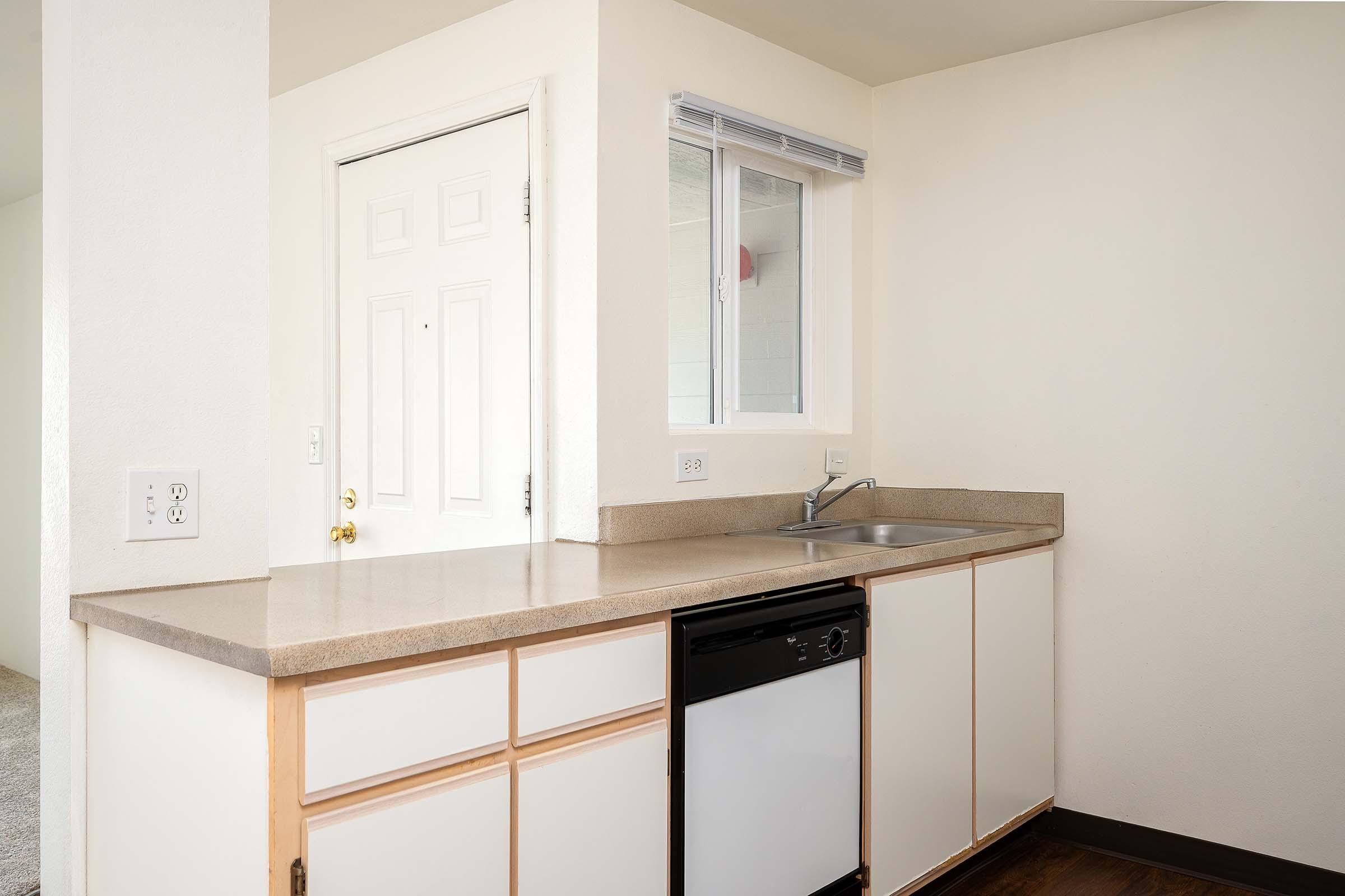 A clean and modern kitchen area featuring a sink, a dishwasher, and light-colored cabinetry. The space is well-lit, with a window above the sink. A white door can be seen in the background, leading to another room. The counter has a smooth finish, and the flooring is dark.