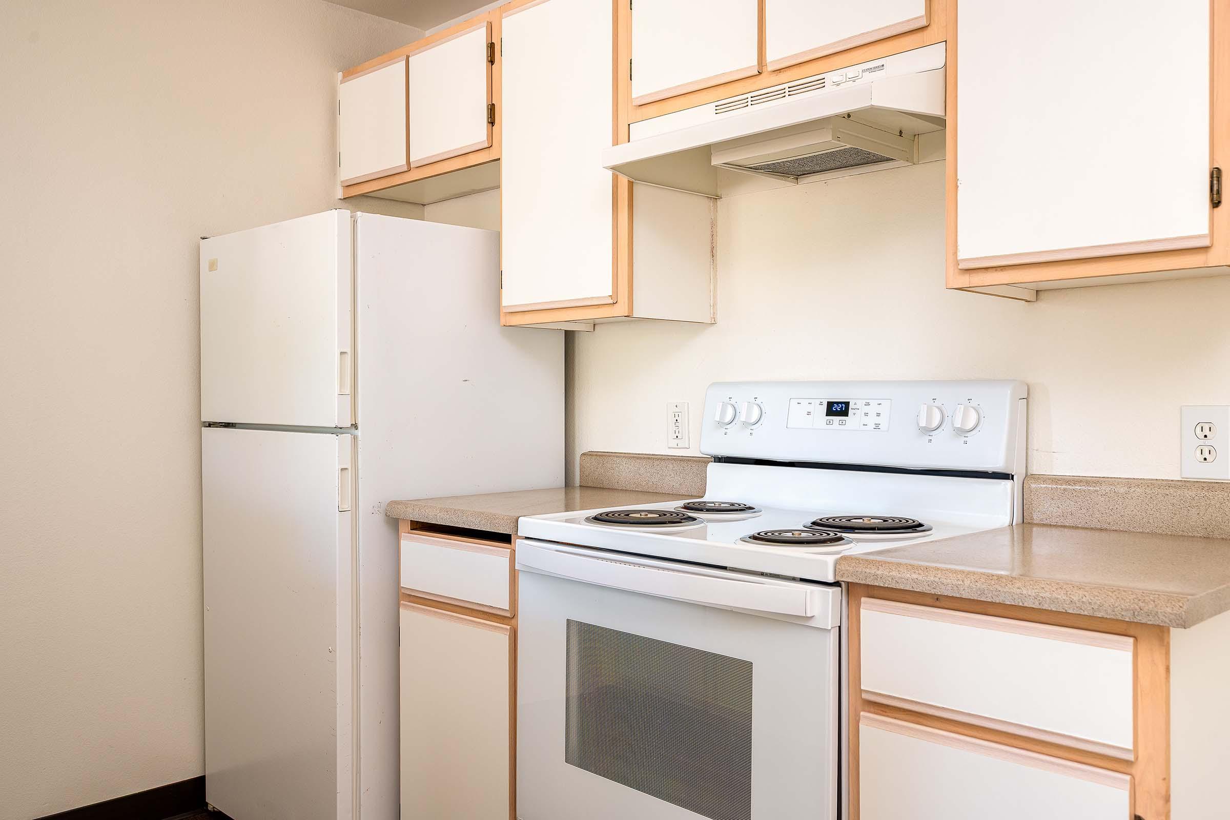 A simple kitchen featuring a white refrigerator, a white stove with four burners, and beige cabinets. The countertops are light-colored, and the walls are plain. The overall design is functional and minimalistic, suitable for basic cooking and storage needs.